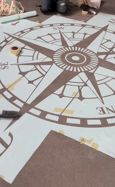 Large floor map of a compass with directions marked, laying flat on the ground, surrounded by people working on it.