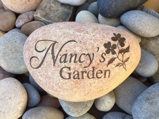 A large pinkish stone with the words "Nancy's Garden" and a flower graphic engraved on it, surrounded by smaller gray, white, and dark stones.