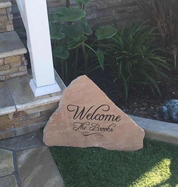 Welcome stone with the text "Welcome The Brooks" placed on a small patch of grass near a stone pathway, plants, and a wooden fence.