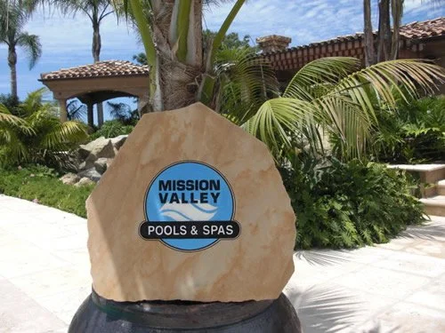Sign for Mission Valley Pools & Spas in a tropical outdoor setting with palm trees and a thatched-roof structure in the background.