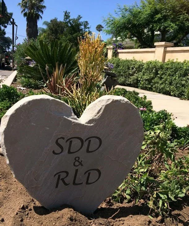 A heart-shaped stone with the inscription 'SDD & RLD' in a garden with various plants and a sidewalk in the background.