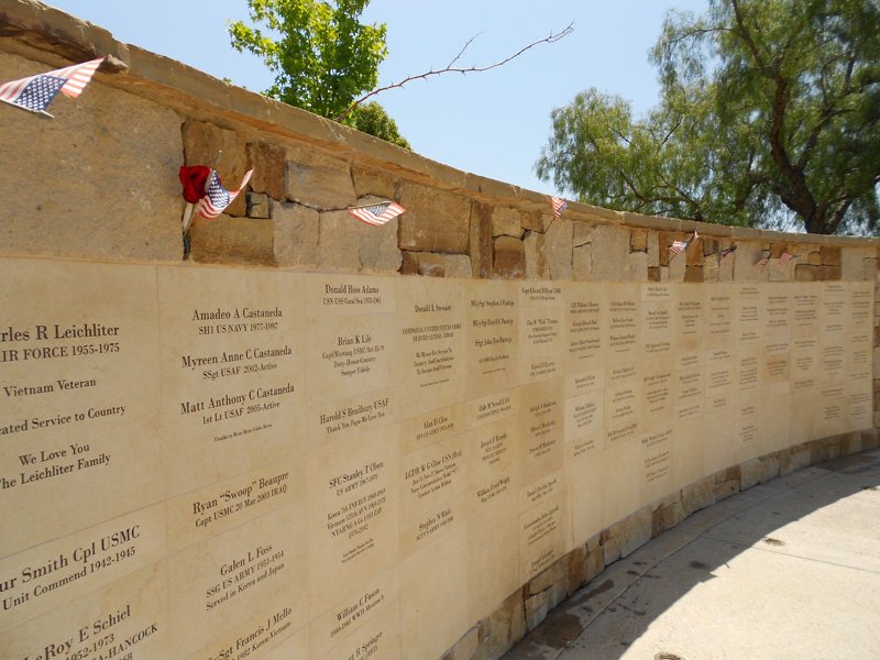 A memorial wall displaying engraved names of military veterans, decorated with small American flags along the top, set against a backdrop of trees and a blue sky.