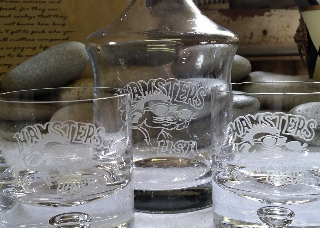 A glass decanter and two glasses with 'WAMSTERS USA' and a rat wearing sunglasses etched on them, placed on a surface with rocks and a handwritten note in the background.