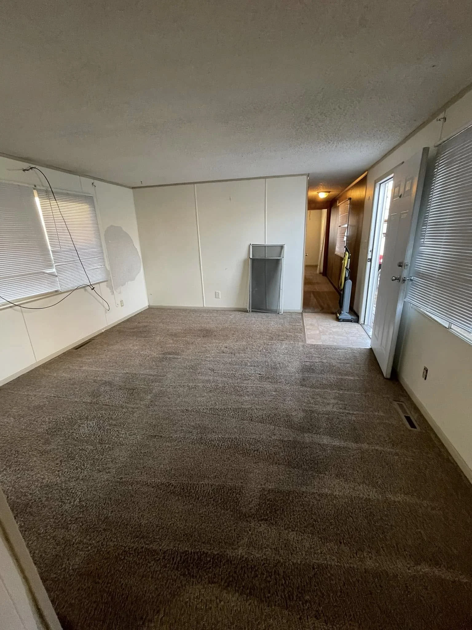Empty living room with brown carpet, white walls, two windows with blinds, and a hallway door. There is a metal shelf and vacuum cleaner near the entrance.