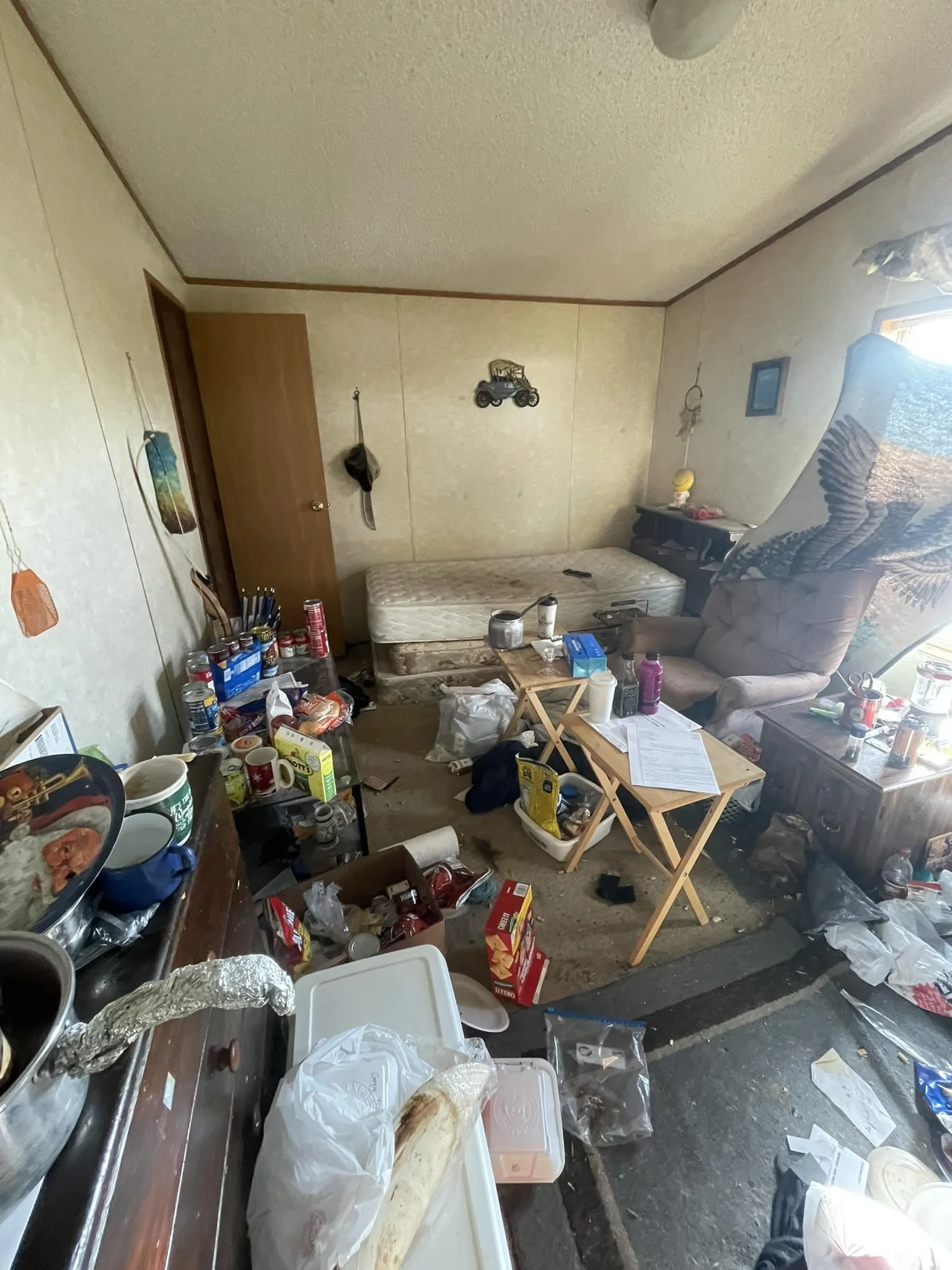 Messy living room with cluttered table, scattered trash, and an unmade mattress. There's a recliner and a window with blinds.