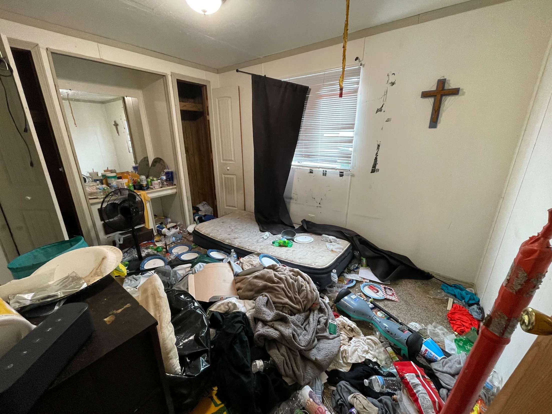A cluttered and messy bedroom with an unmade mattress on the floor, scattered trash, clothes, bottles, and food containers. The room has a small window with blinds, a mirror on the wall, and a religious cross hanging. There is an open closet and a fan on the floor.