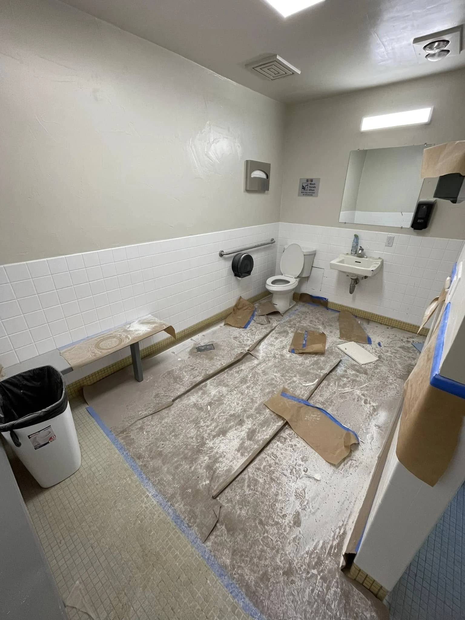 A bathroom under renovation with tiles removed from the floor, covering the concrete subfloor. Fixtures include a toilet, a small sink, a mirror, a soap dispenser, and various construction materials and paper covering the floor.