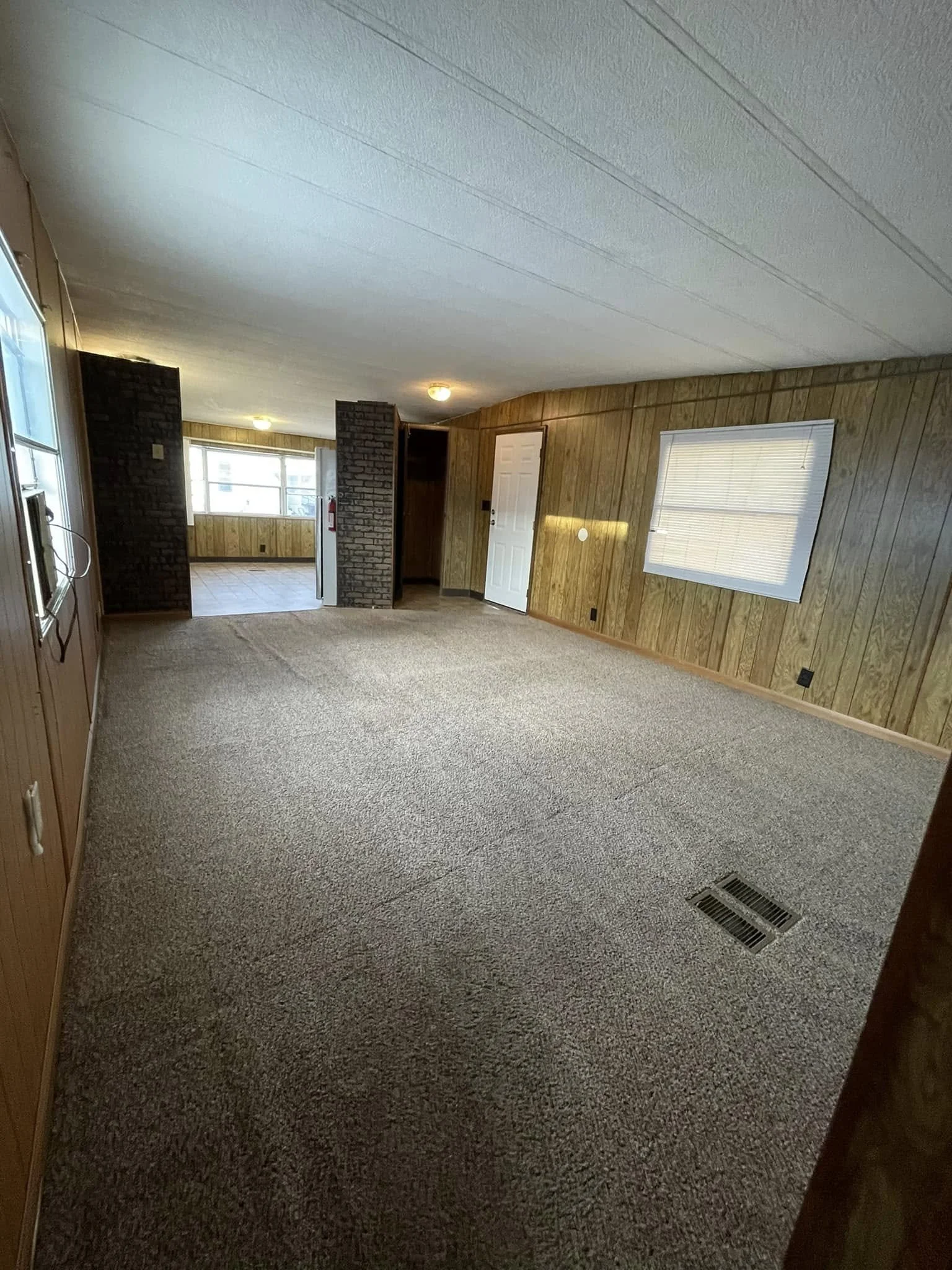 Empty living room with wood panel walls, carpet flooring, and a window with blinds, leading to a sunlit area with large windows.