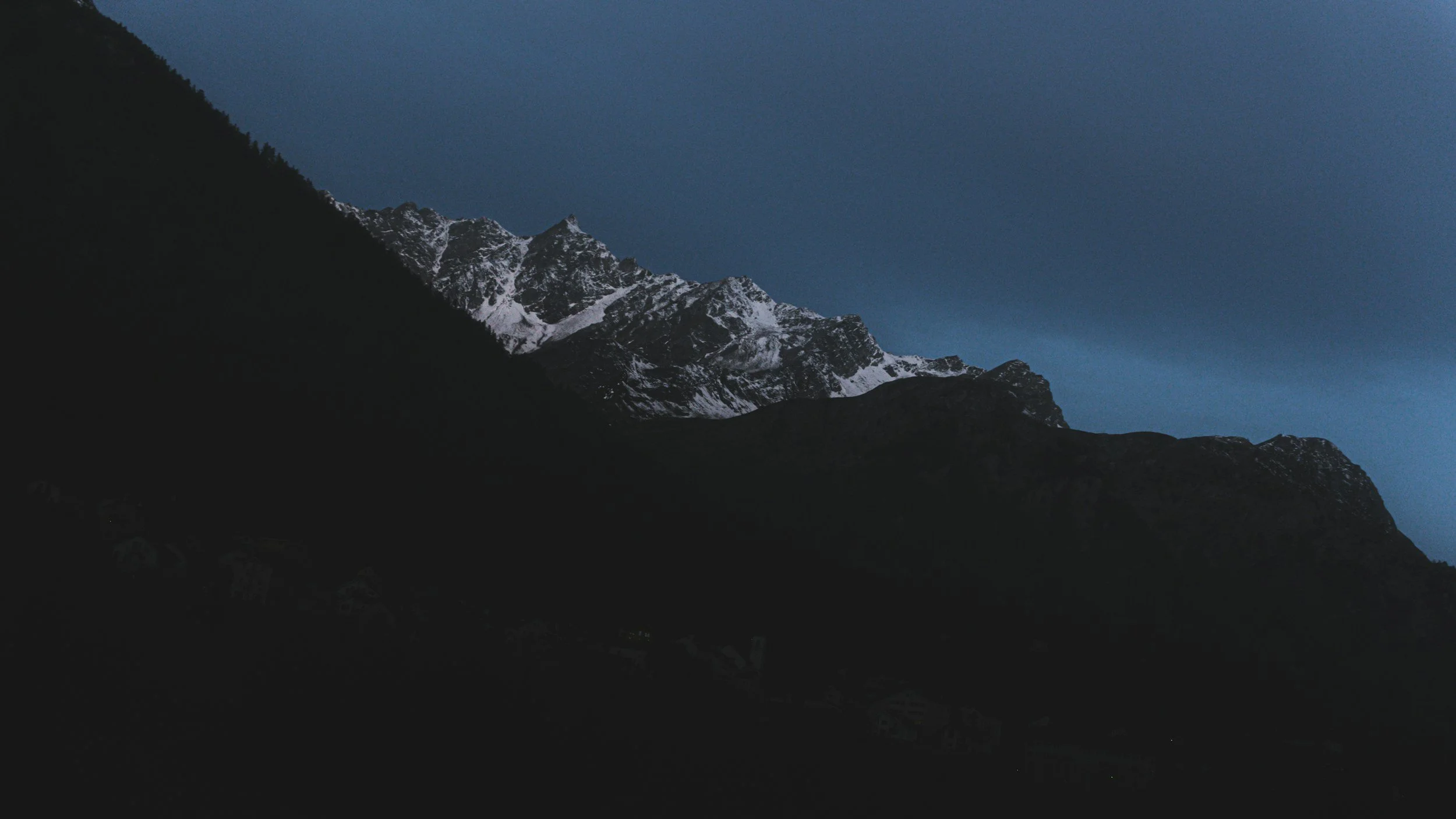 Nighttime mountain landscape with snow-capped peaks and dark hills in foreground, deep blue sky overhead.