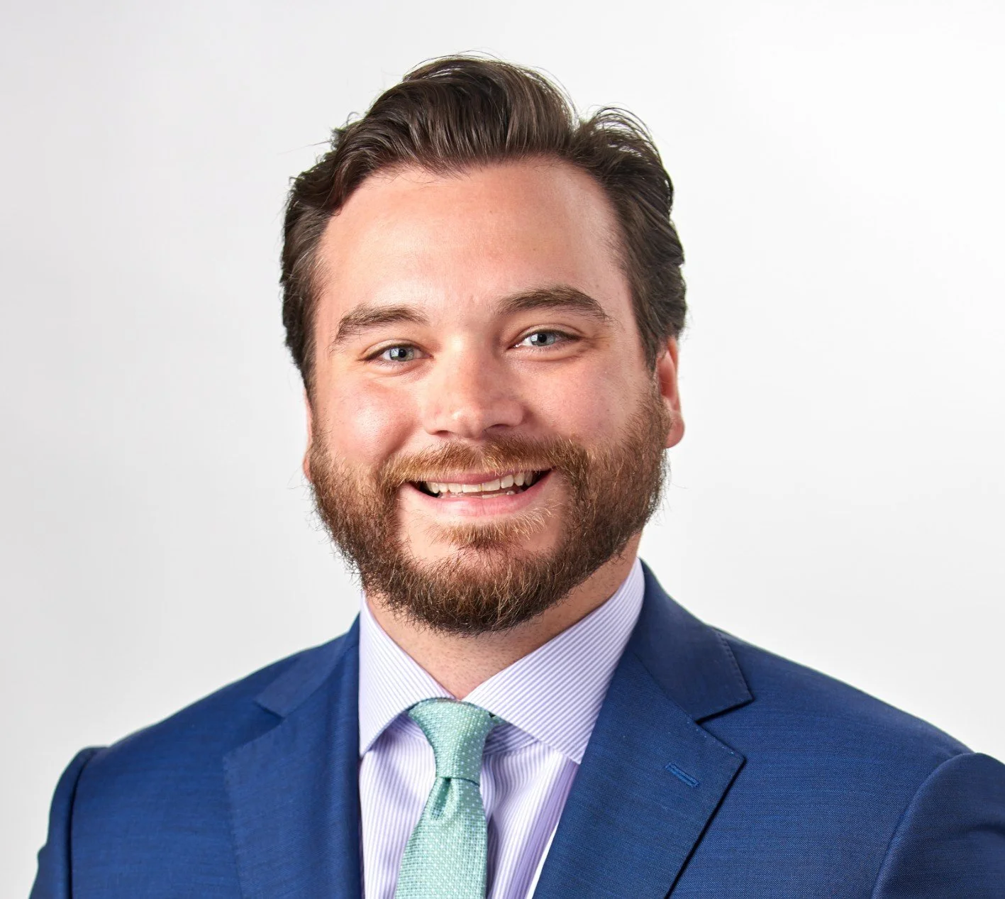 Adult man with a beard and mustache smiling, wearing a blue suit, light purple shirt, and light green tie, with a white background.