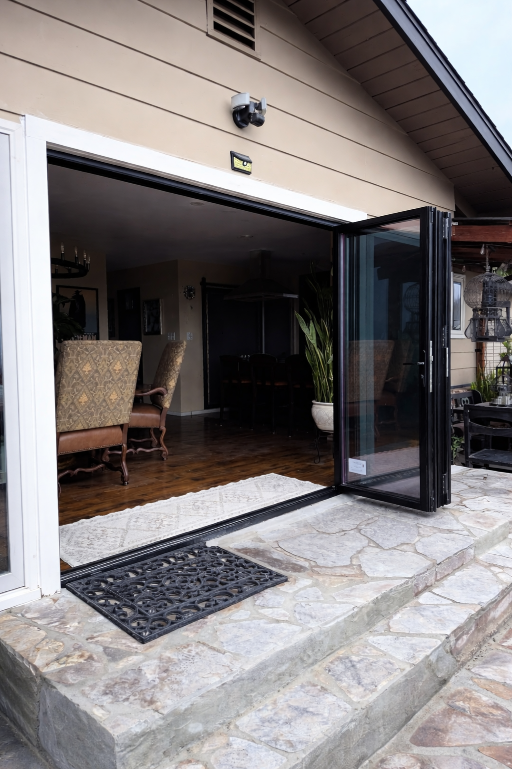Open glass sliding door leading from outdoor stone patio into a living room with wooden flooring, armchairs, and potted plants.