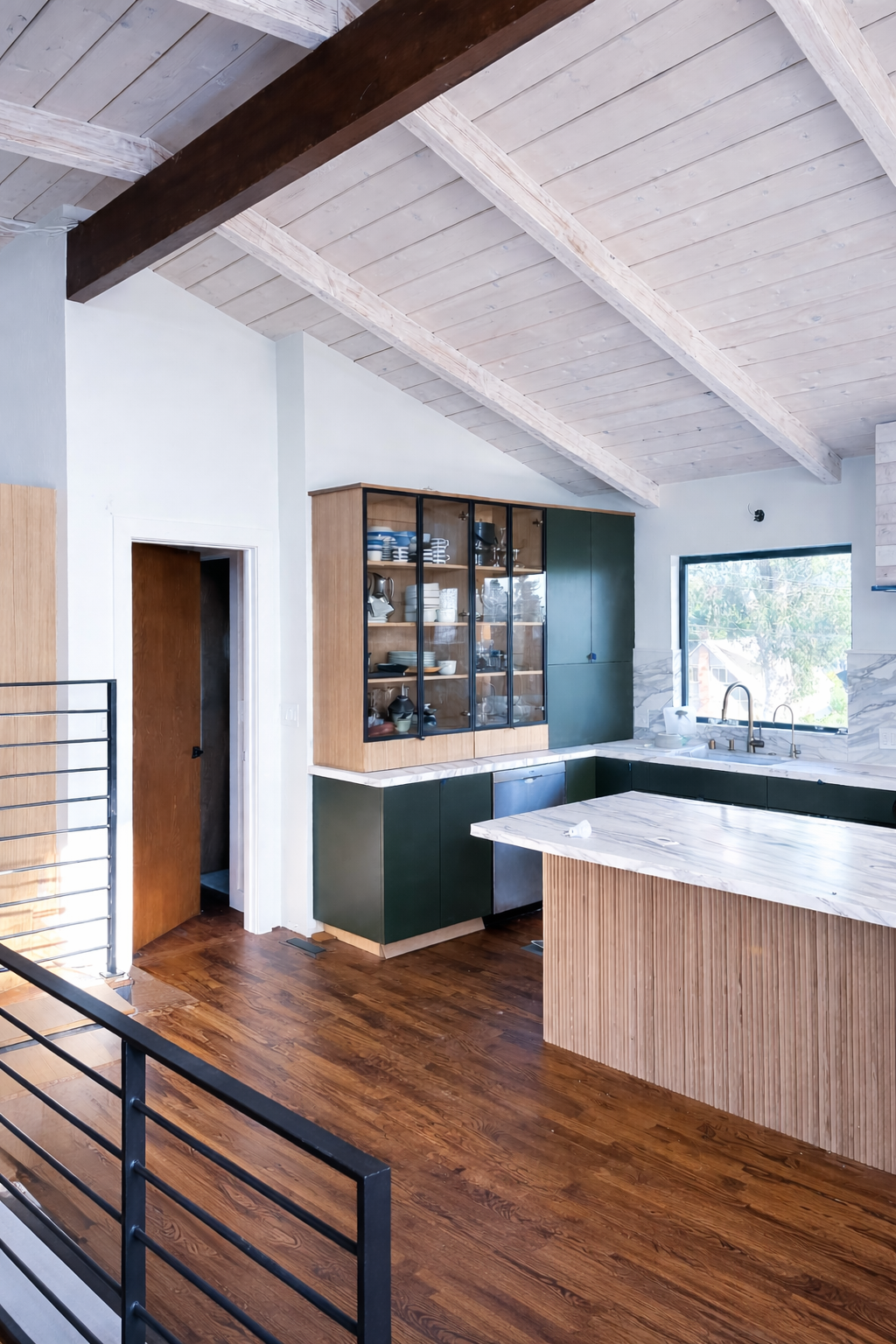 Modern kitchen with wooden cabinets, marble countertops, and a large window, featuring a vaulted ceiling with exposed wooden beams and hardwood flooring.