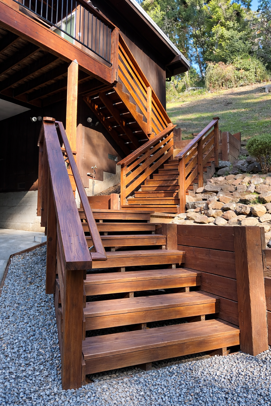 Wooden outdoor stairs and railing leading to an elevated house with a balcony, surrounded by rocks and trees.