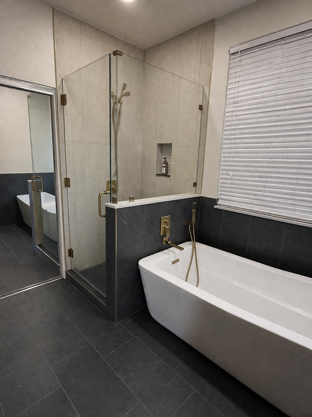 A modern bathroom features a white bathtub with a gold faucet and fixtures, a glass-enclosed shower with a handheld showerhead, and a window with closed white blinds, all with neutral tile walls and dark floor tiles.