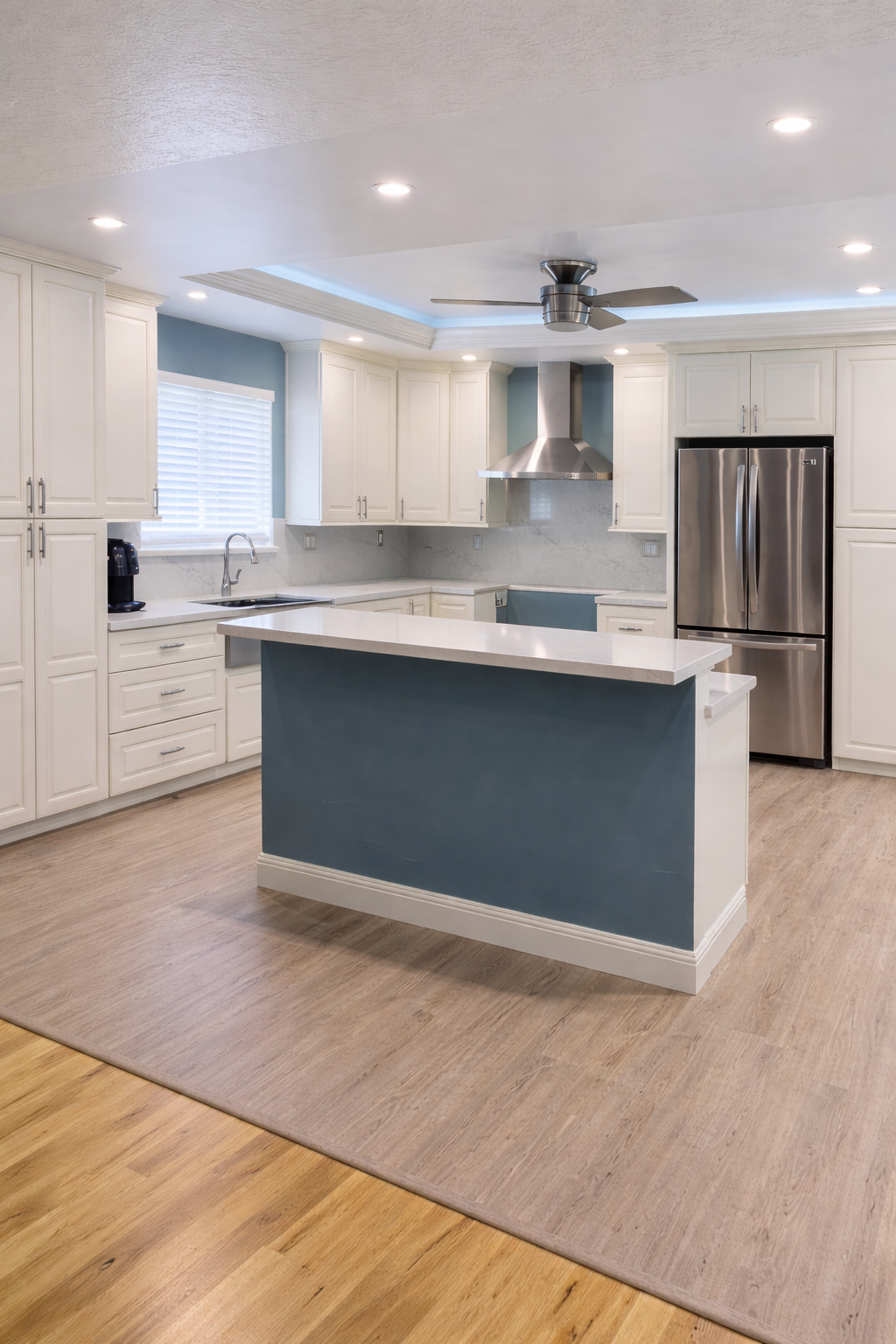 Modern kitchen with white cabinets, stainless steel refrigerator and range hood, center island with blue base, light-colored hardwood and vinyl flooring, ceiling fan, window with blinds, and recessed lighting.
