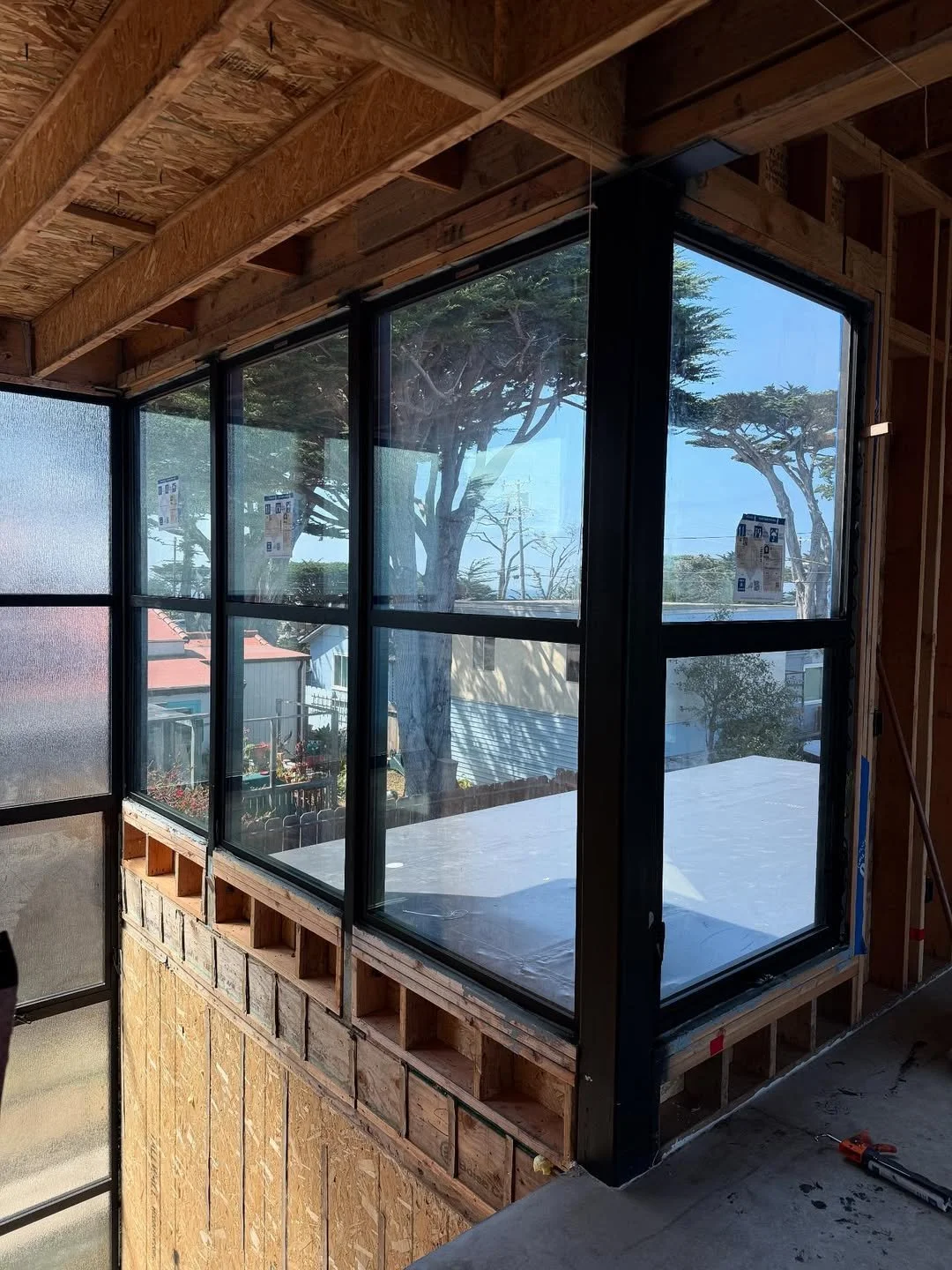 Interior view of a building under construction with large black-framed windows, wooden framing, and a view of trees and neighboring houses outside.