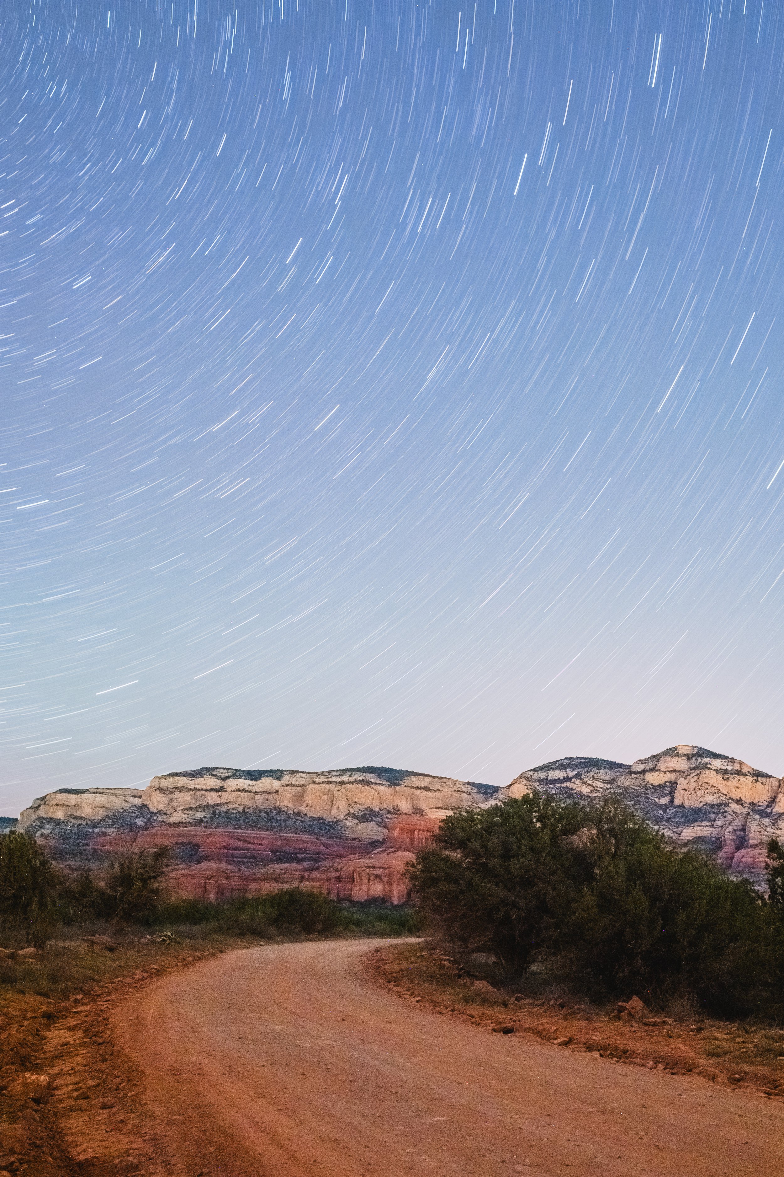 Star trails over desert landscape with rocky cliffs and a winding dirt road.
