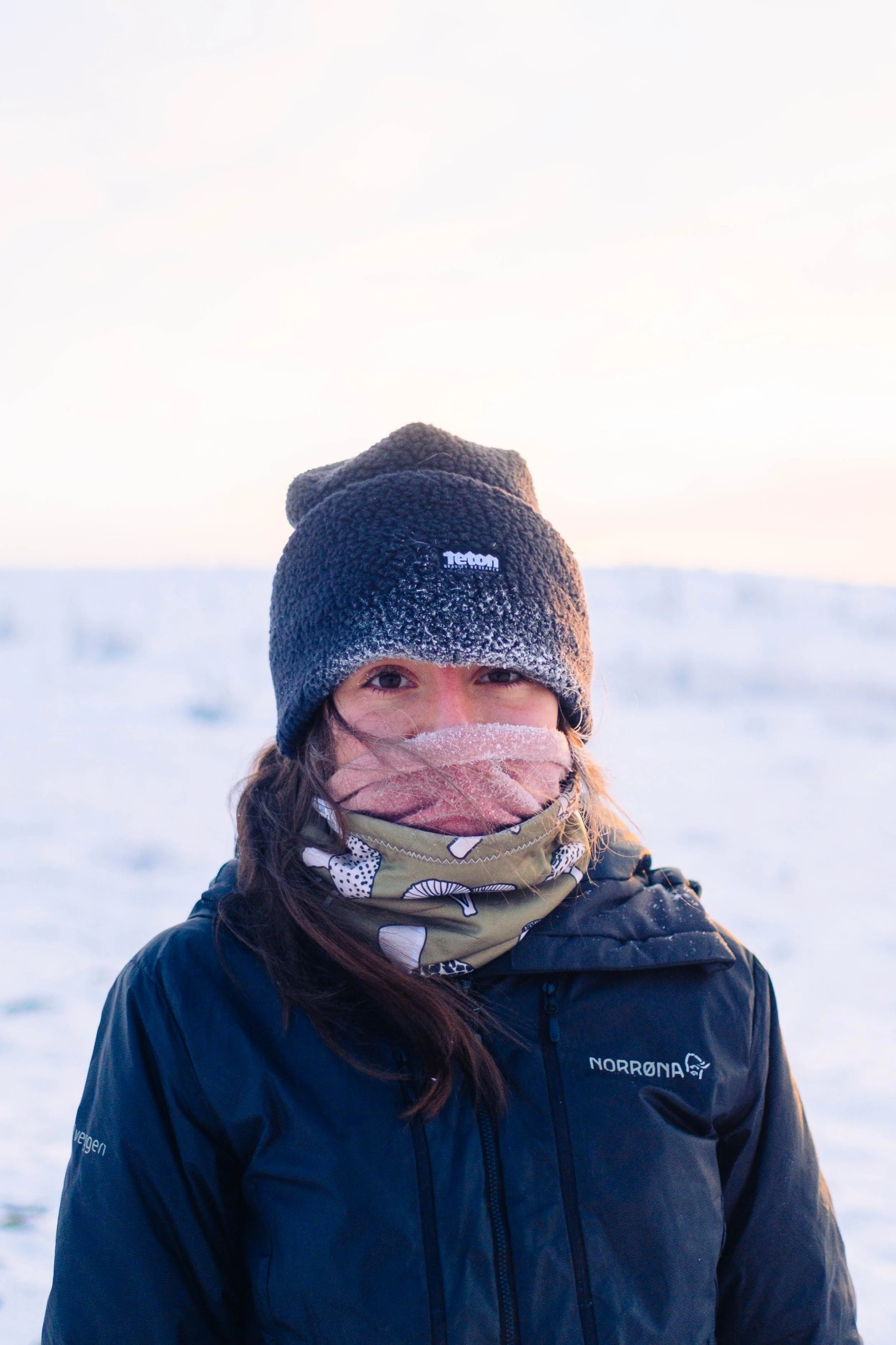 Close-up of a woman dressed for cold weather in a beanie, scarf, and winter coat with snow in the background.