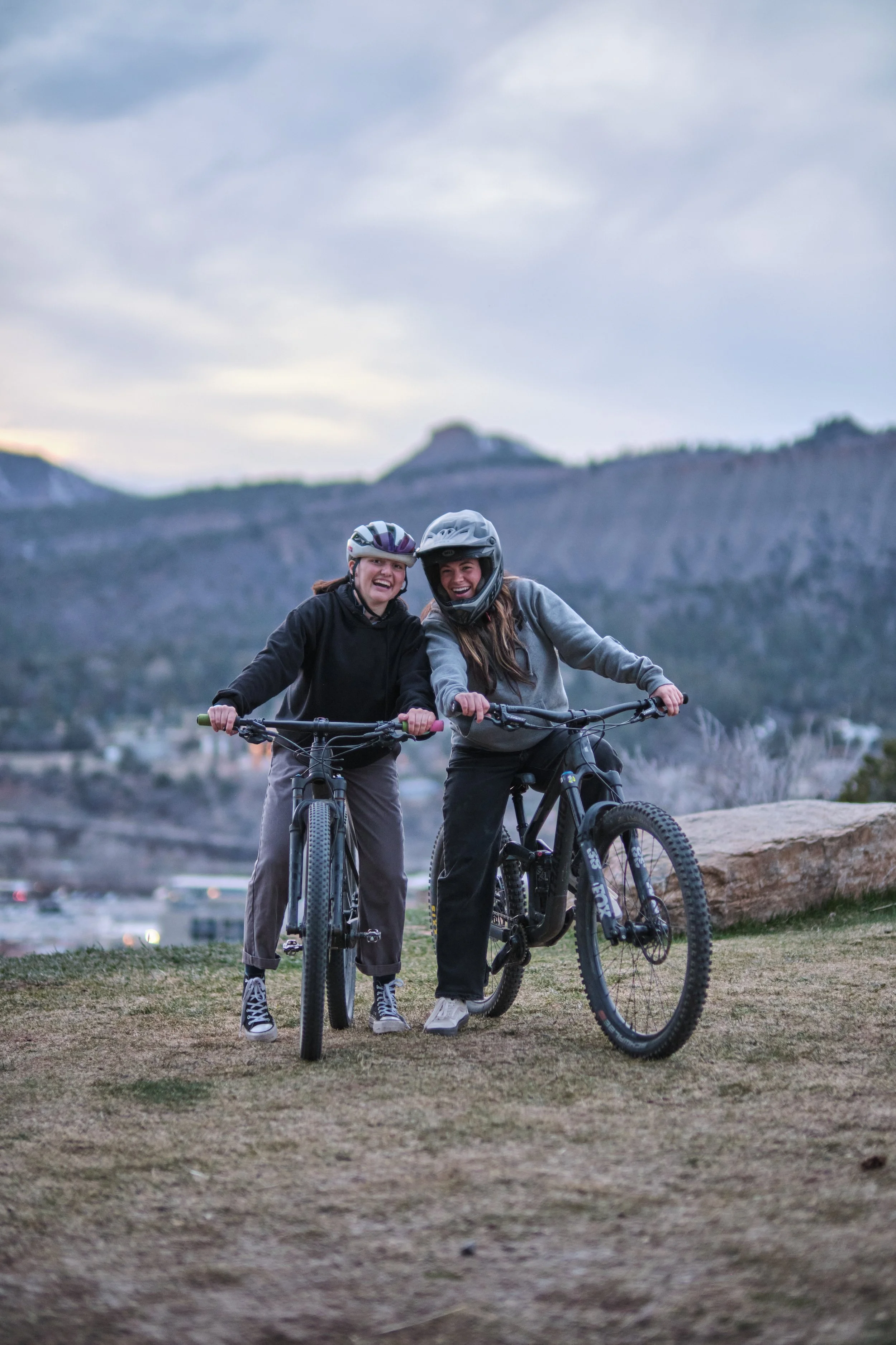 Two women wearing helmets riding mountain bikes outdoors, smiling and enjoying a scenic landscape with mountains in the background during dusk.