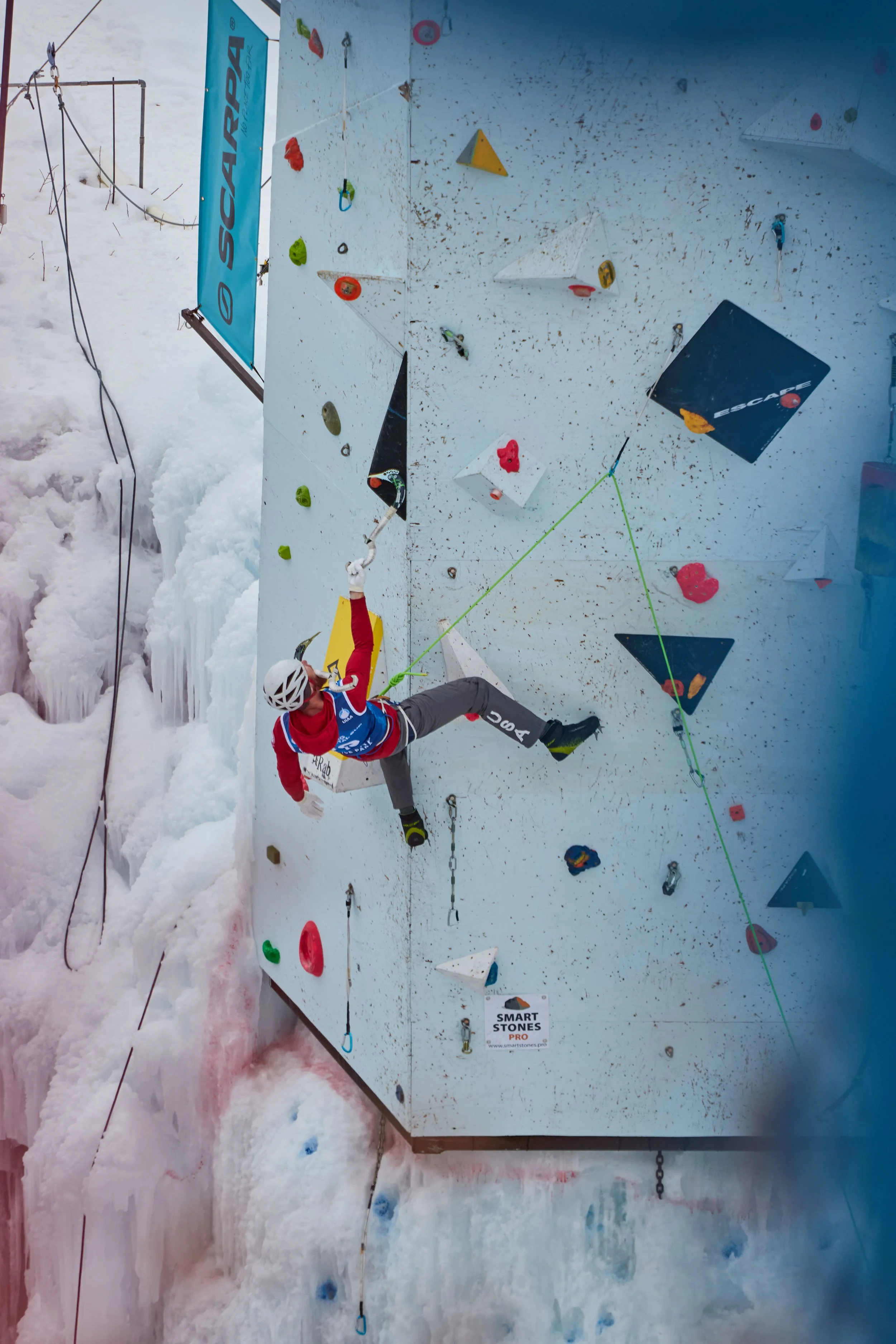 An indoor ice climbing wall with colorful holds and a climber wearing a helmet and harness ascending. The wall has various routes marked by different colored holds and is surrounded by snow at the base. Ice climber at Ouray ice festival