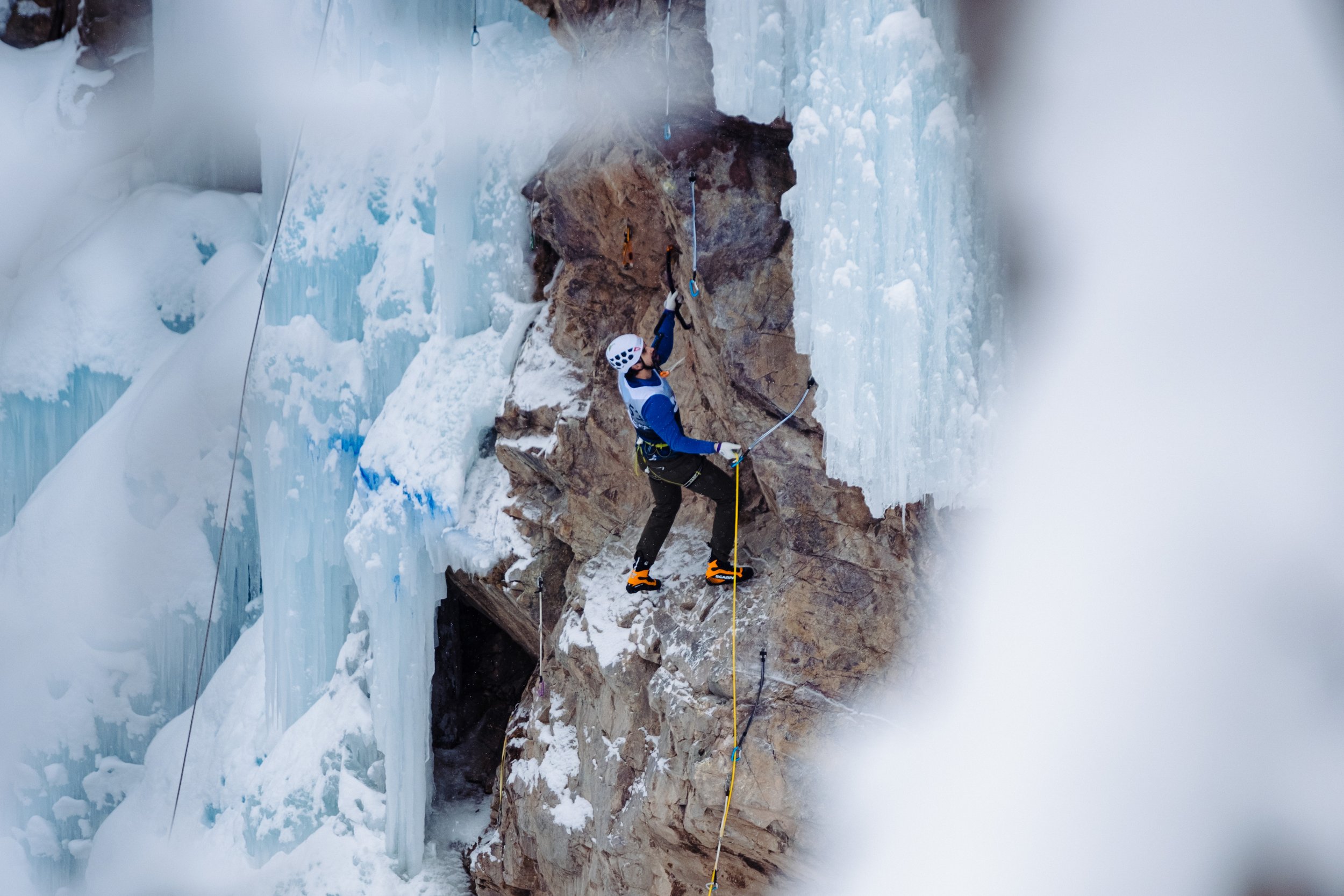 A person ice climbing on a frozen waterfall with safety gear, including a helmet, harness, and climbing tools, surrounded by snow and ice.