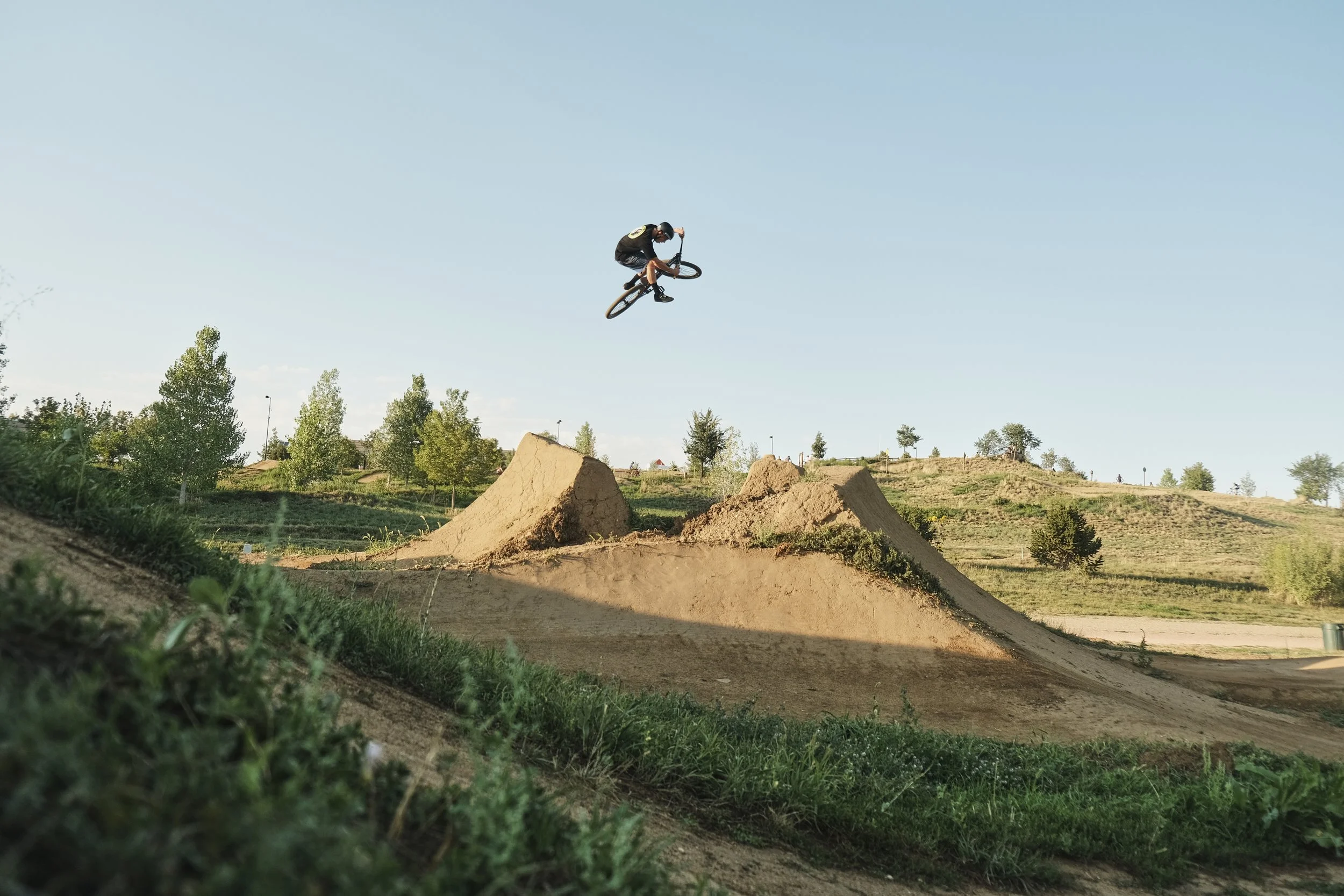 A person performing a stunt on a dirt jump bicycle in a dirt park, mid-air above a dirt mound with trees and a clear blue sky in the background. Valmont bike park, mountain bike, box, dirt jump, cycling photographer.