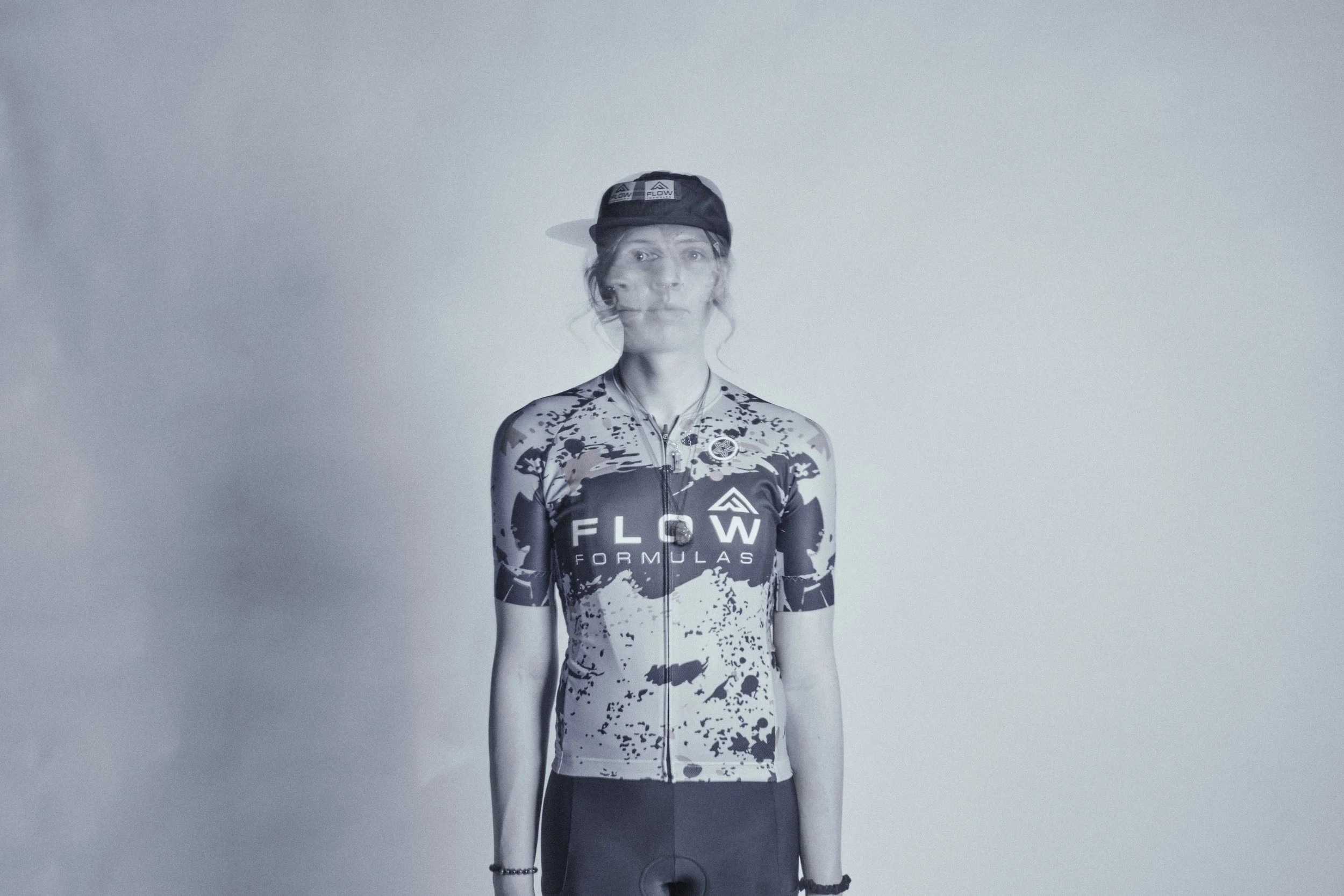 A woman in cycling gear with a black and white splatter pattern jersey that has 'FLOW FORMULAS' written on it, standing against a plain background with a neutral expression, wearing a black cap. cyclist, photoshoot, durango cycling