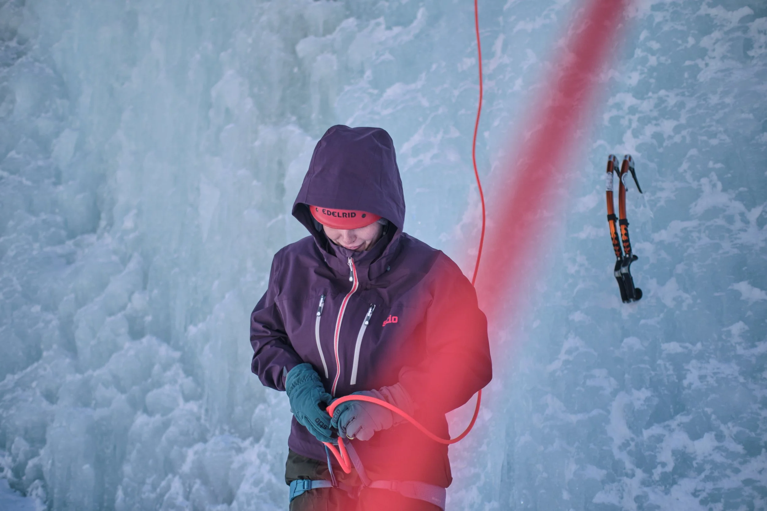 A person dressed in winter clothing, including a purple jacket and a beanie, standing in front of an icy glacier, holding a climbing rope connected to ice climbing tools hanging on their side.
