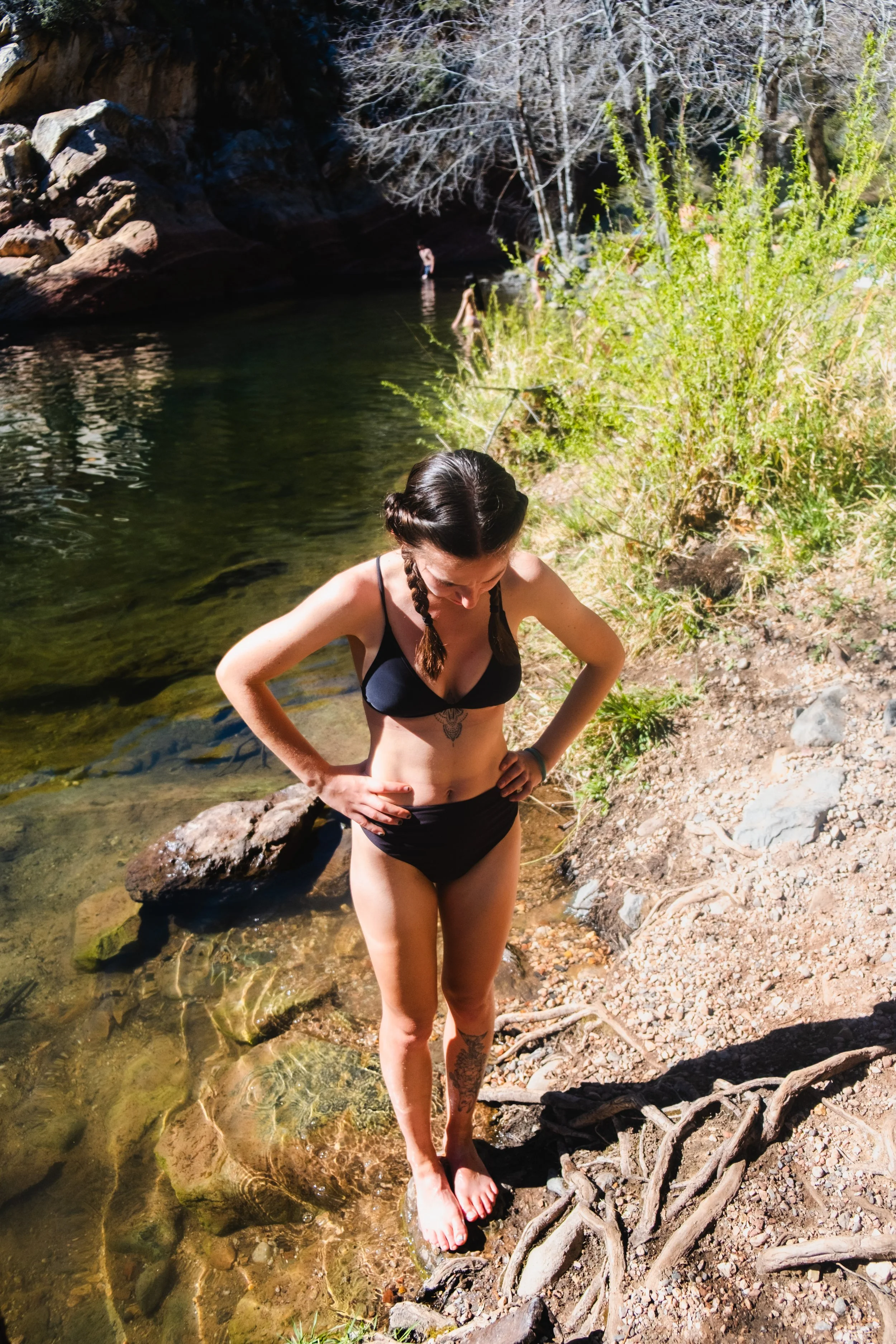 A woman in a black bikini standing in shallow water at the edge of a rocky creek or river, with several people swimming and wading in the background and trees with dry branches and green foliage along the bank.