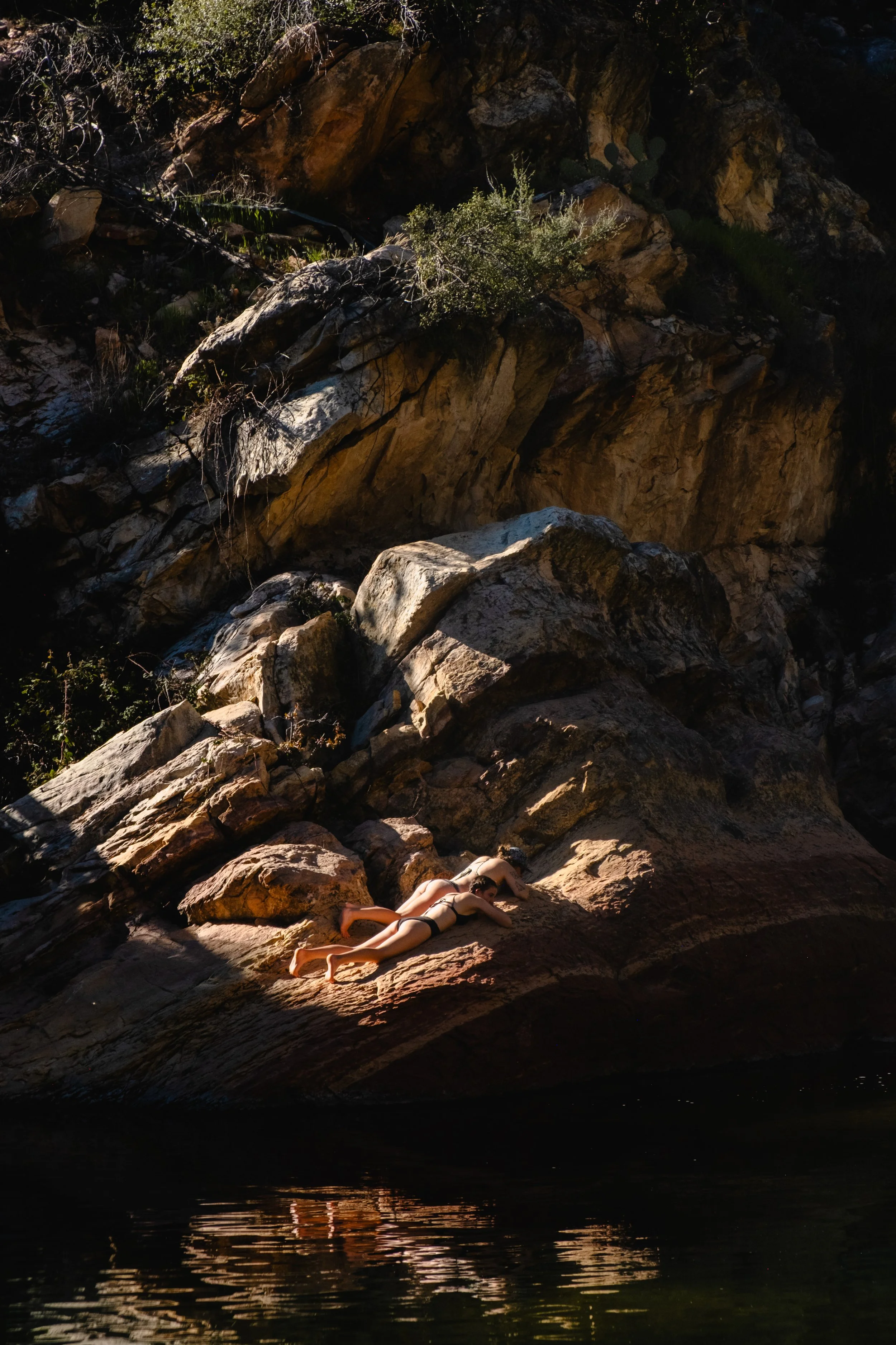 A woman sunbathing on a rocky riverbank surrounded by large rocks and cliffs.