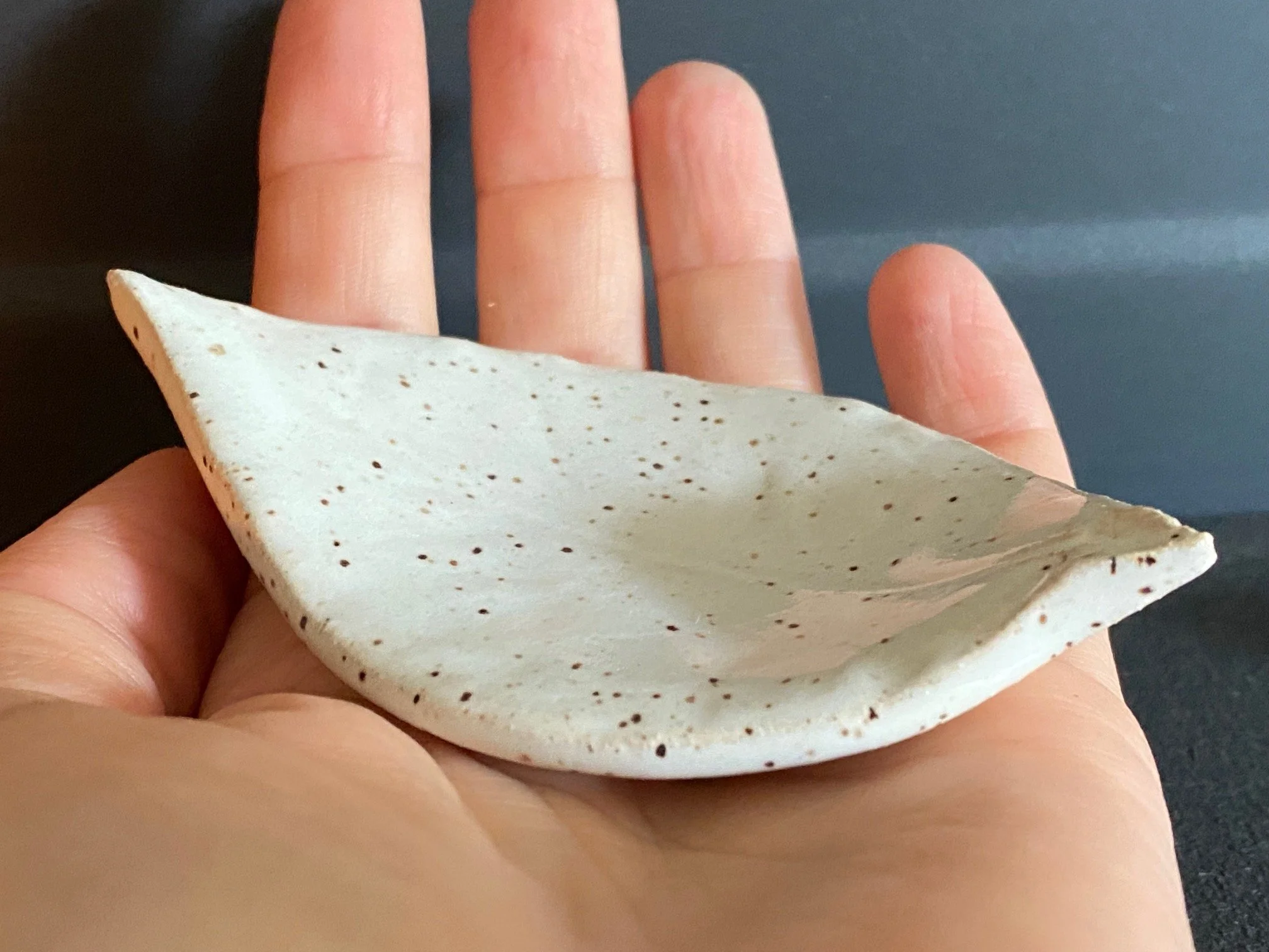 A person's hand holding a white ceramic tea light holder shaped like a elongated leaf against a dark background. Sonia Lesage Ceramics