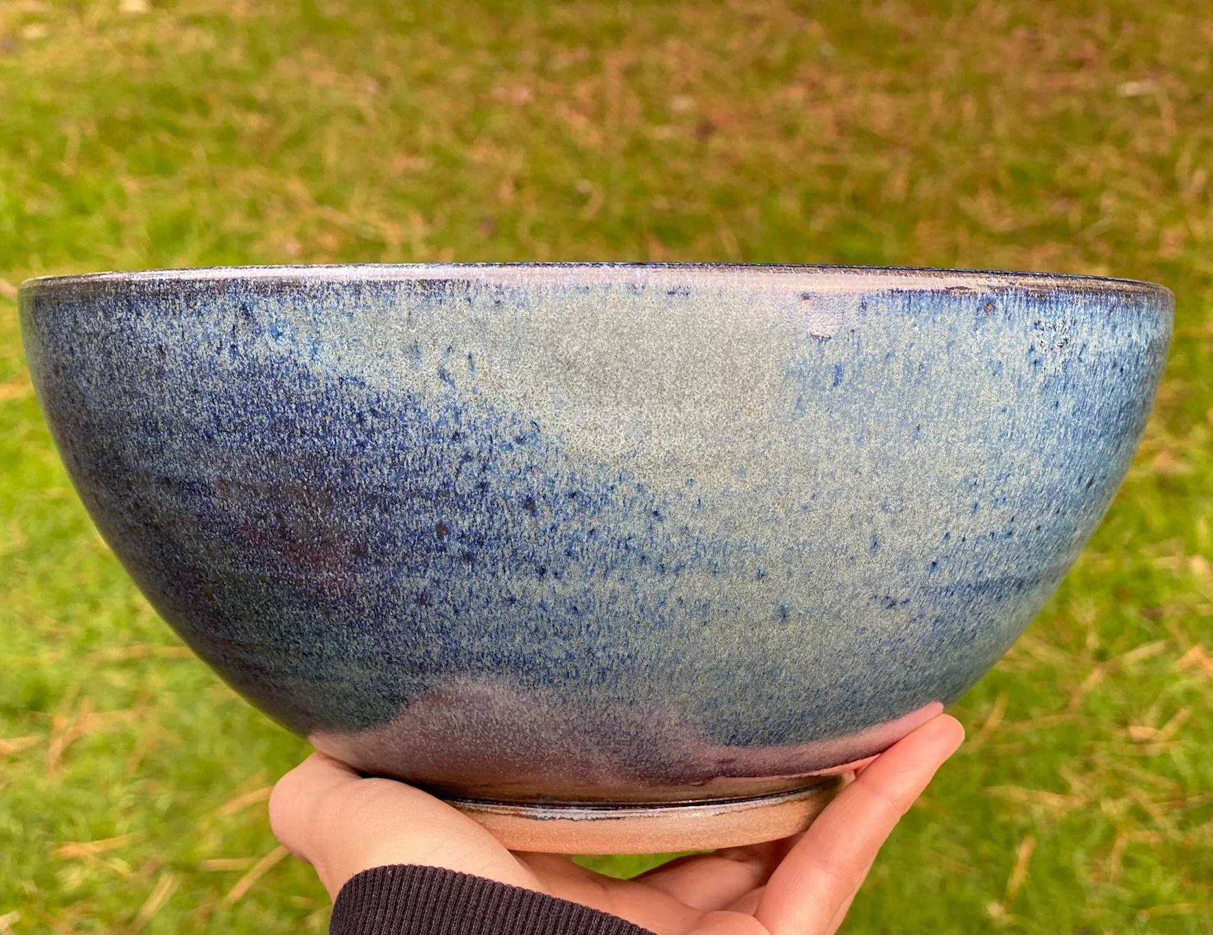 A hand holding a large, round ceramic bowl with a textured, two-toned glaze in shades of blue, set outdoors on grass. Sonia Lesage Ceramics