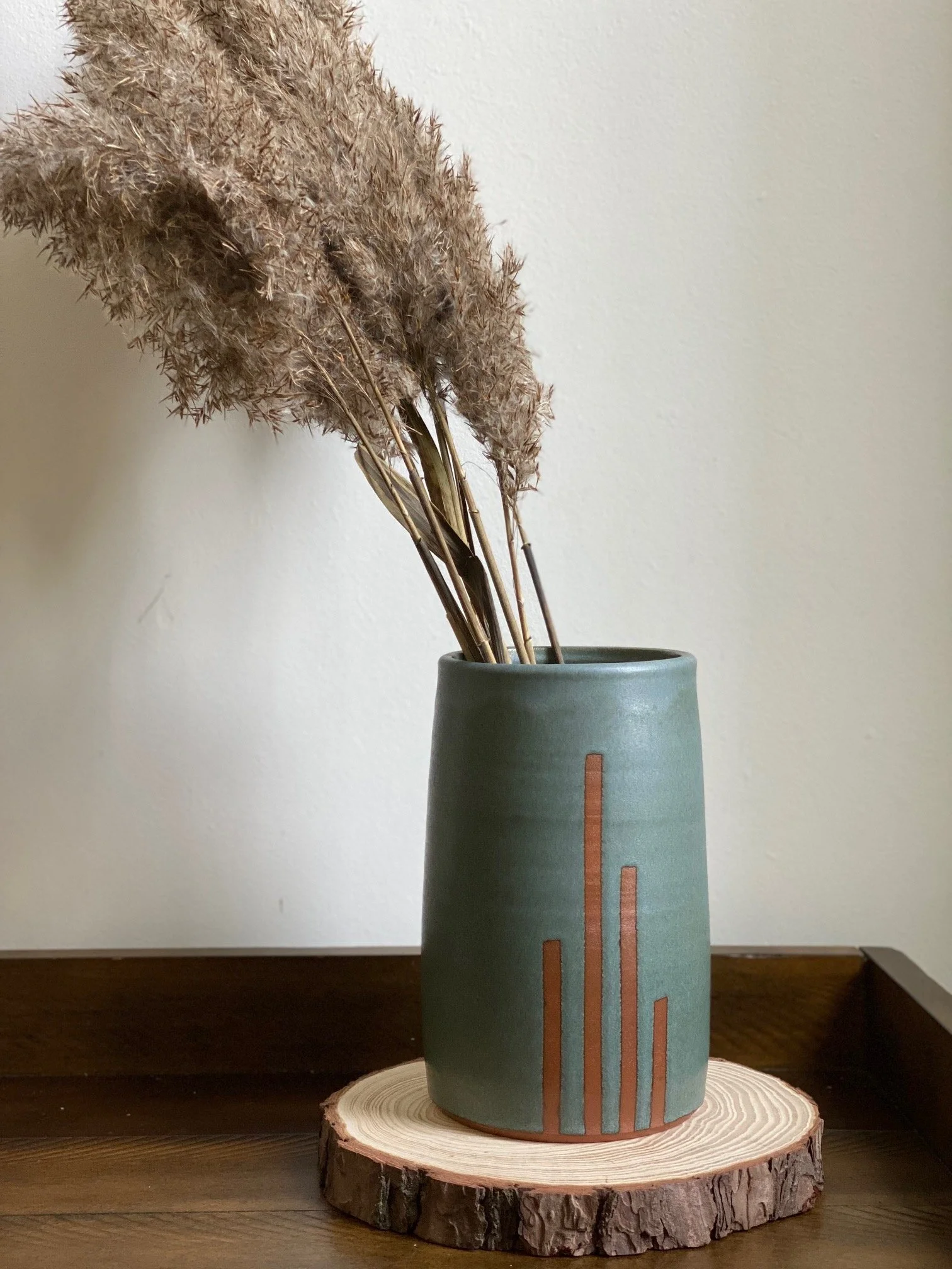 A blue ceramic vase with vertical lines of exposed red clay, holding dried pampas grass, placed on a wood slice on a dark wooden surface against a light-colored wall. Sonia Lesage Ceramics