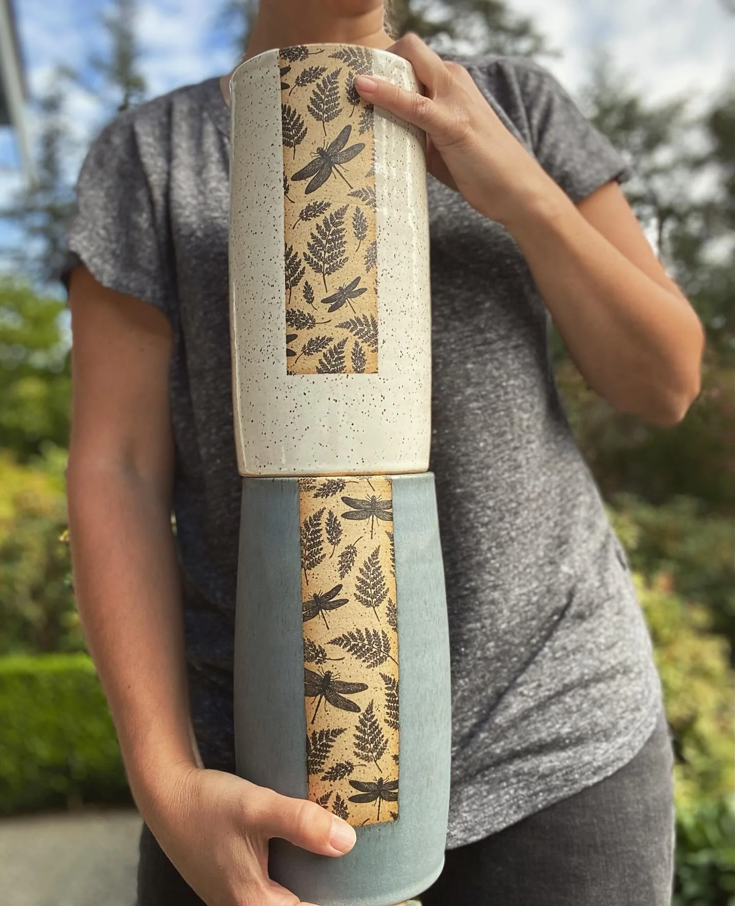 Person holding a tall ceramic vases with botanical and dragonfly patterns, outdoors with trees and sky in the background. Sonia Lesage Ceramics
