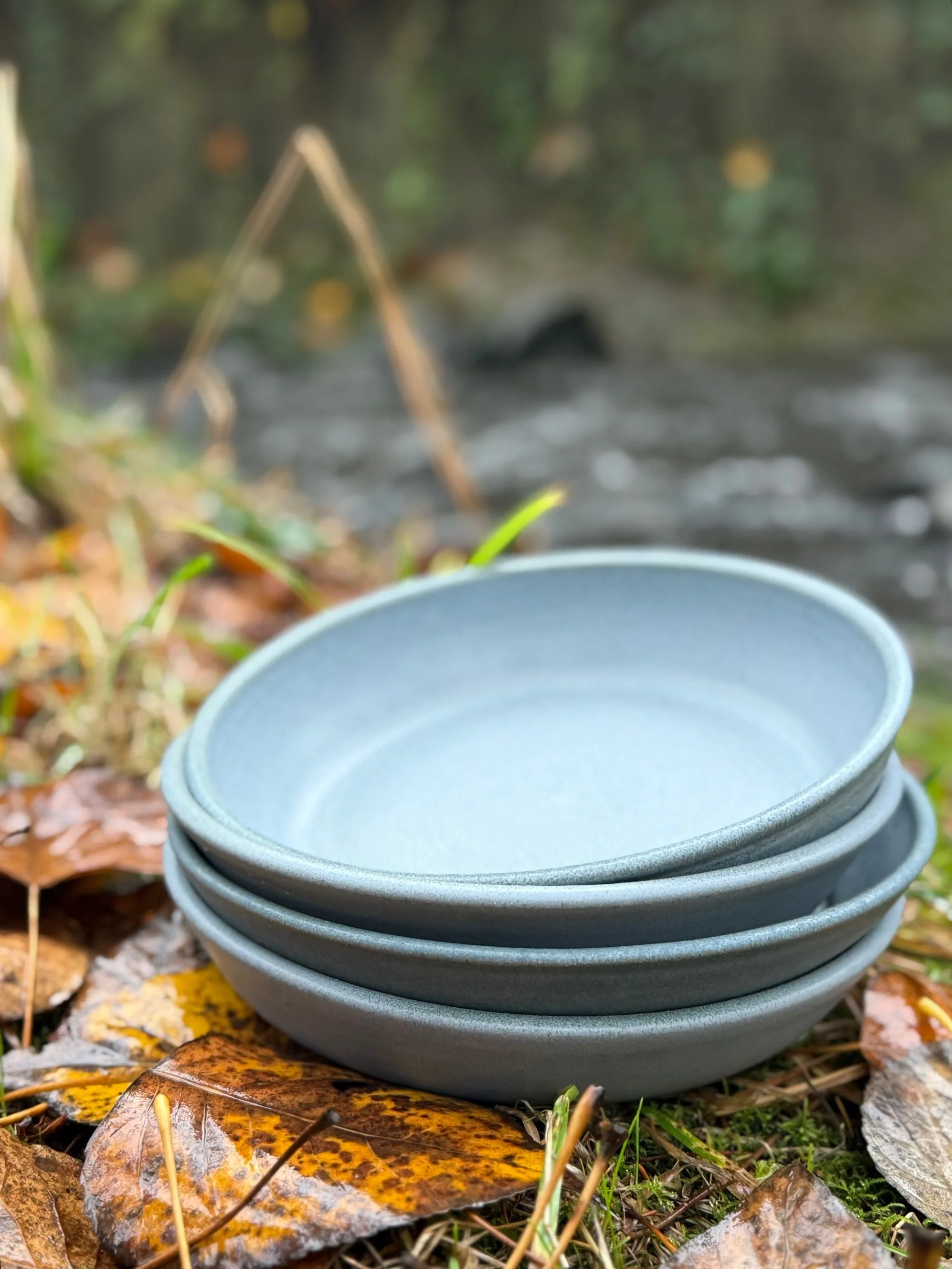 A stack of blue/gray ceramic plates placed on fallen autumn leaves near a riverbank. Sonia Lesage Ceramics