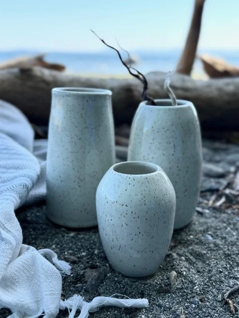 Three speckled white ceramic bud vases on a sandy beach with driftwood and a blurred ocean background. Sonia Lesage Ceramics