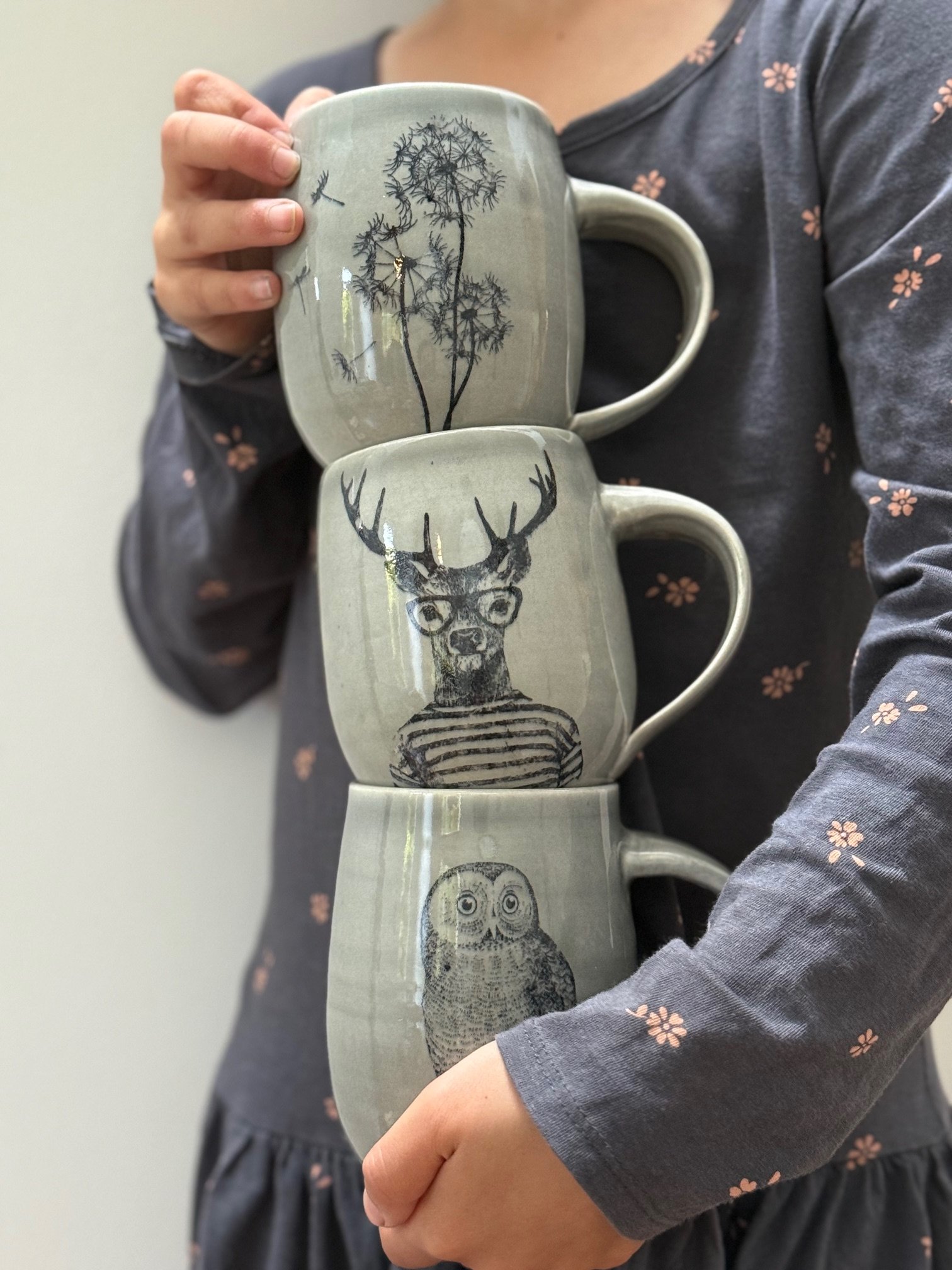 Person holding three stacked ceramic mugs featuring nature-themed silkscreen illustrations of an owl, a deer, and a dandelion plant. Sonia Lesage Ceramics