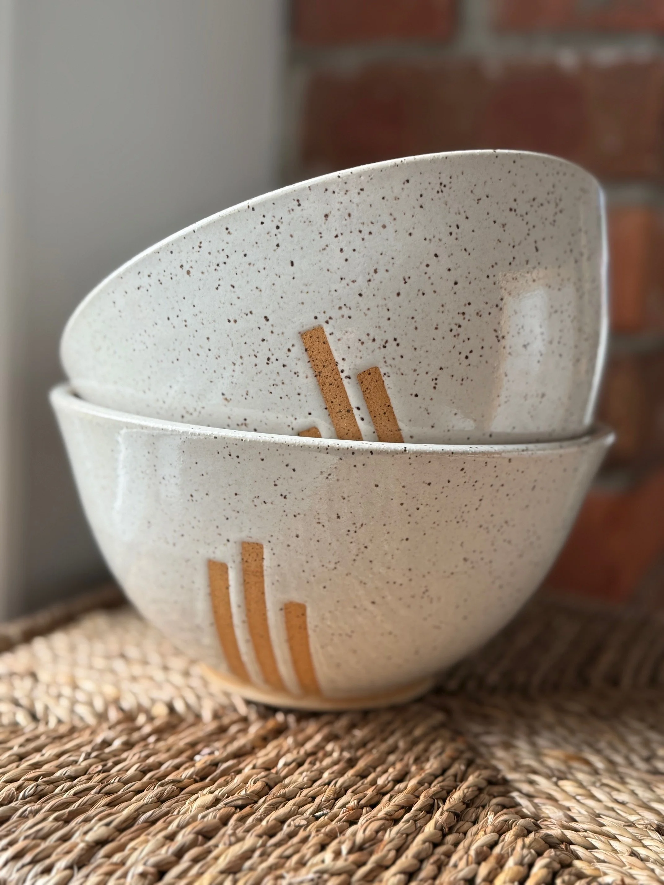 Two stacked speckled white ceramic bowls with geometric lines of exposed clay, placed on woven mat against brick wall background. Sonia Lesage Ceramics