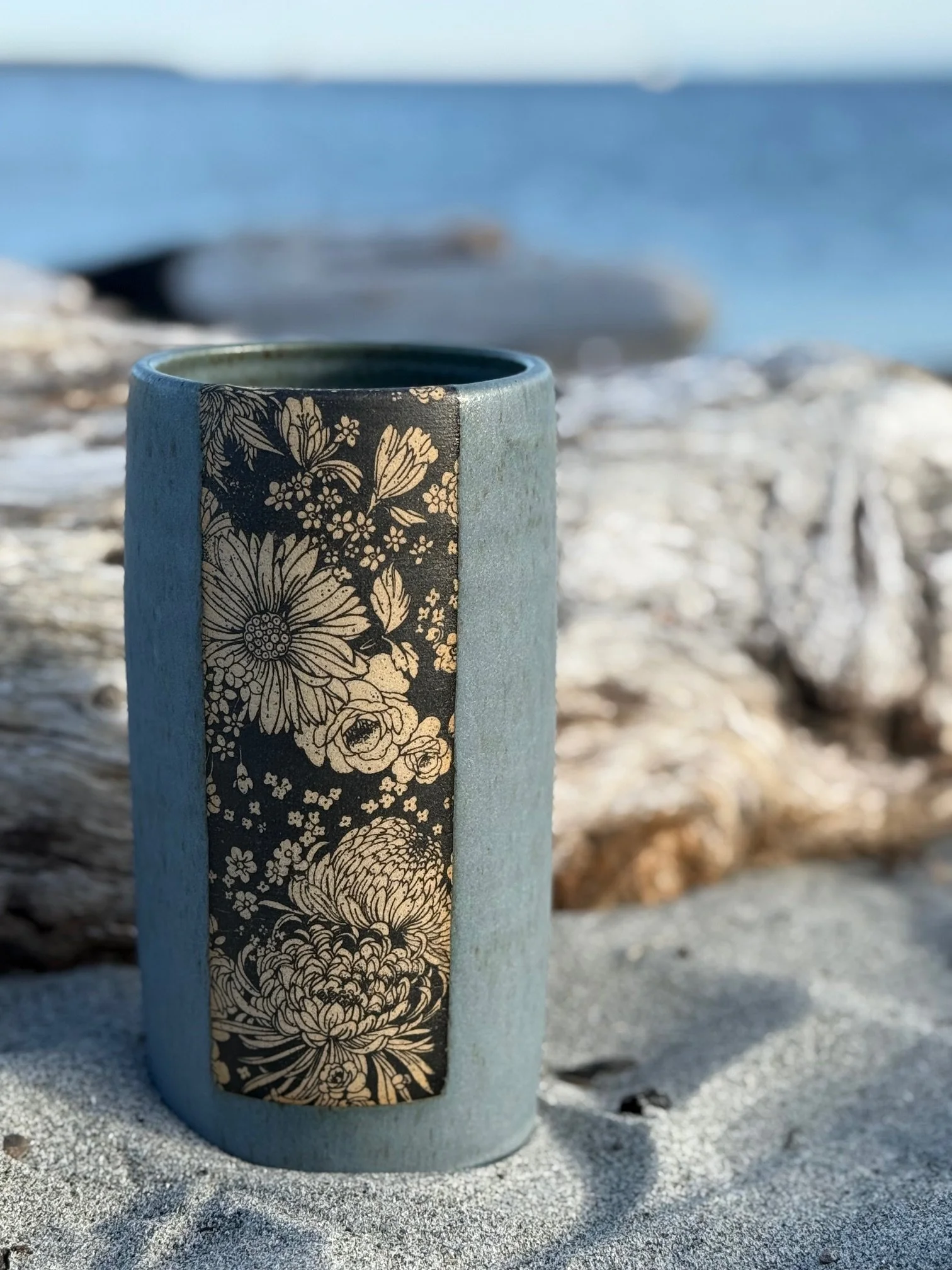 A ceramic vase with floral design on a sandy beach near the ocean, with driftwood and rocks in the background. Sonia Lesage Ceramics