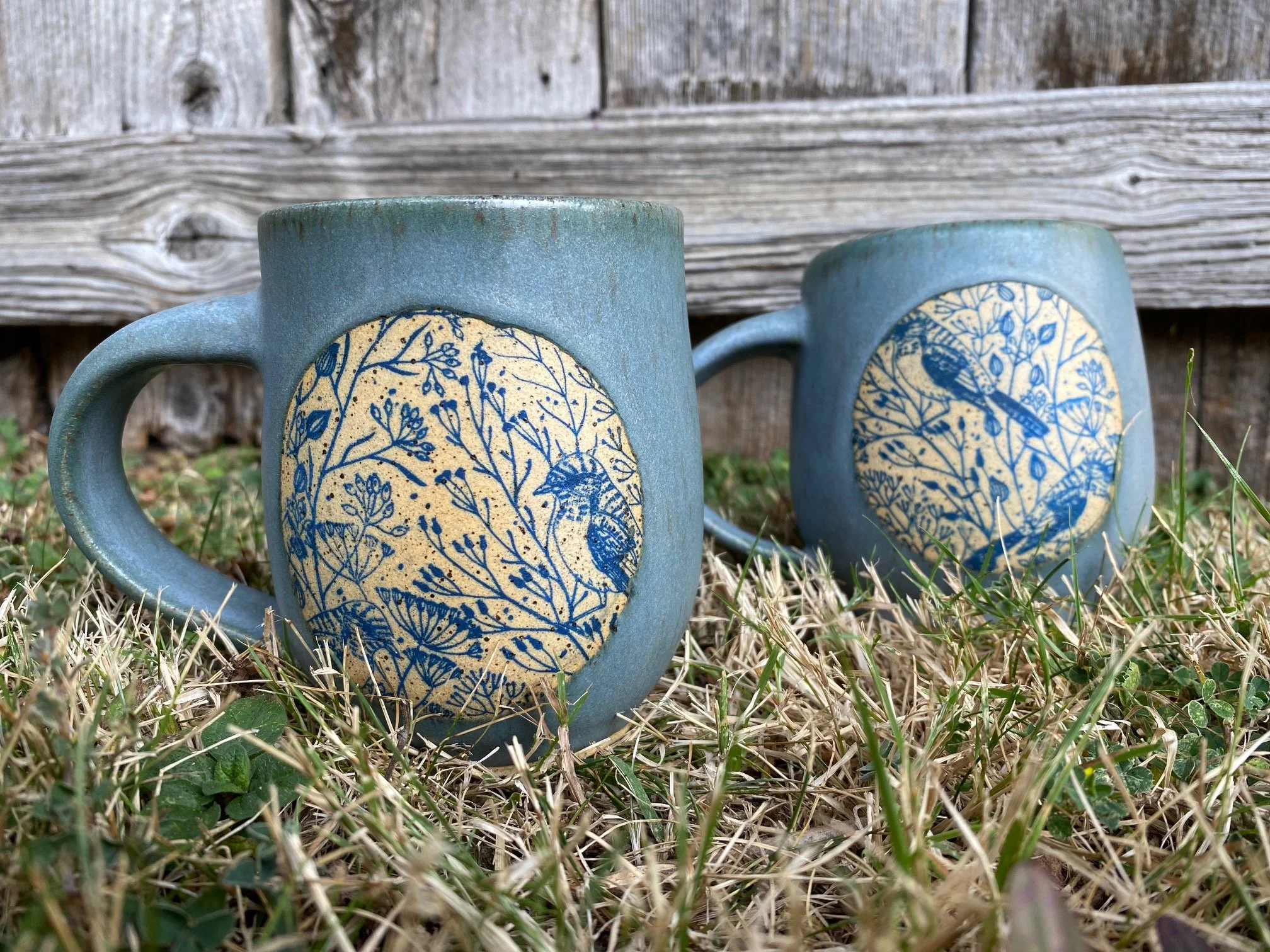 Two ceramic mugs with blue bird and plant designs on them, placed on grass in front of a wooden fence. Sonia Lesage Ceramics