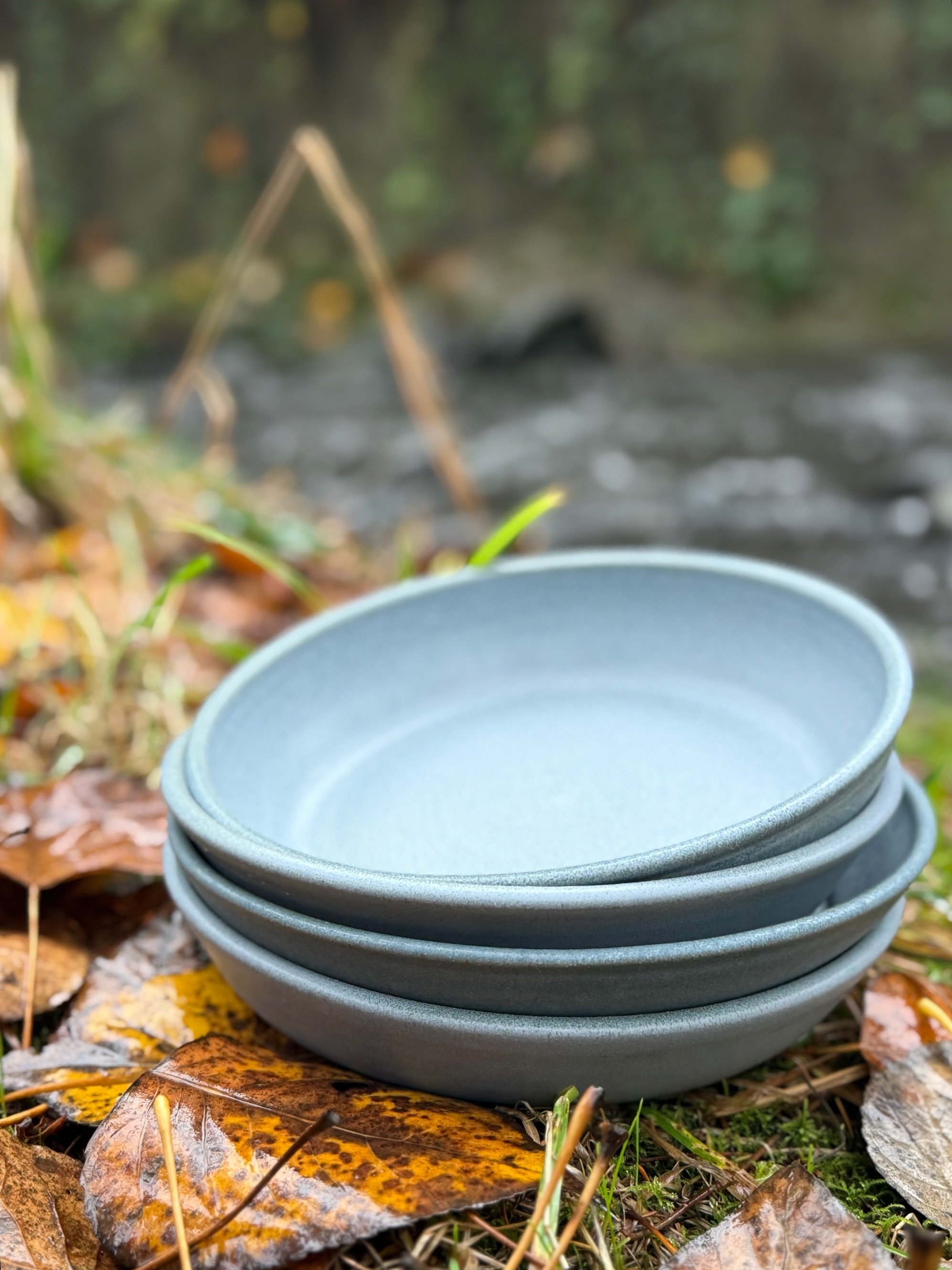 A stack of five gray ceramic plates placed outdoors on fallen autumn leaves near a small stream in a forest. Sonia Lesage Ceramics