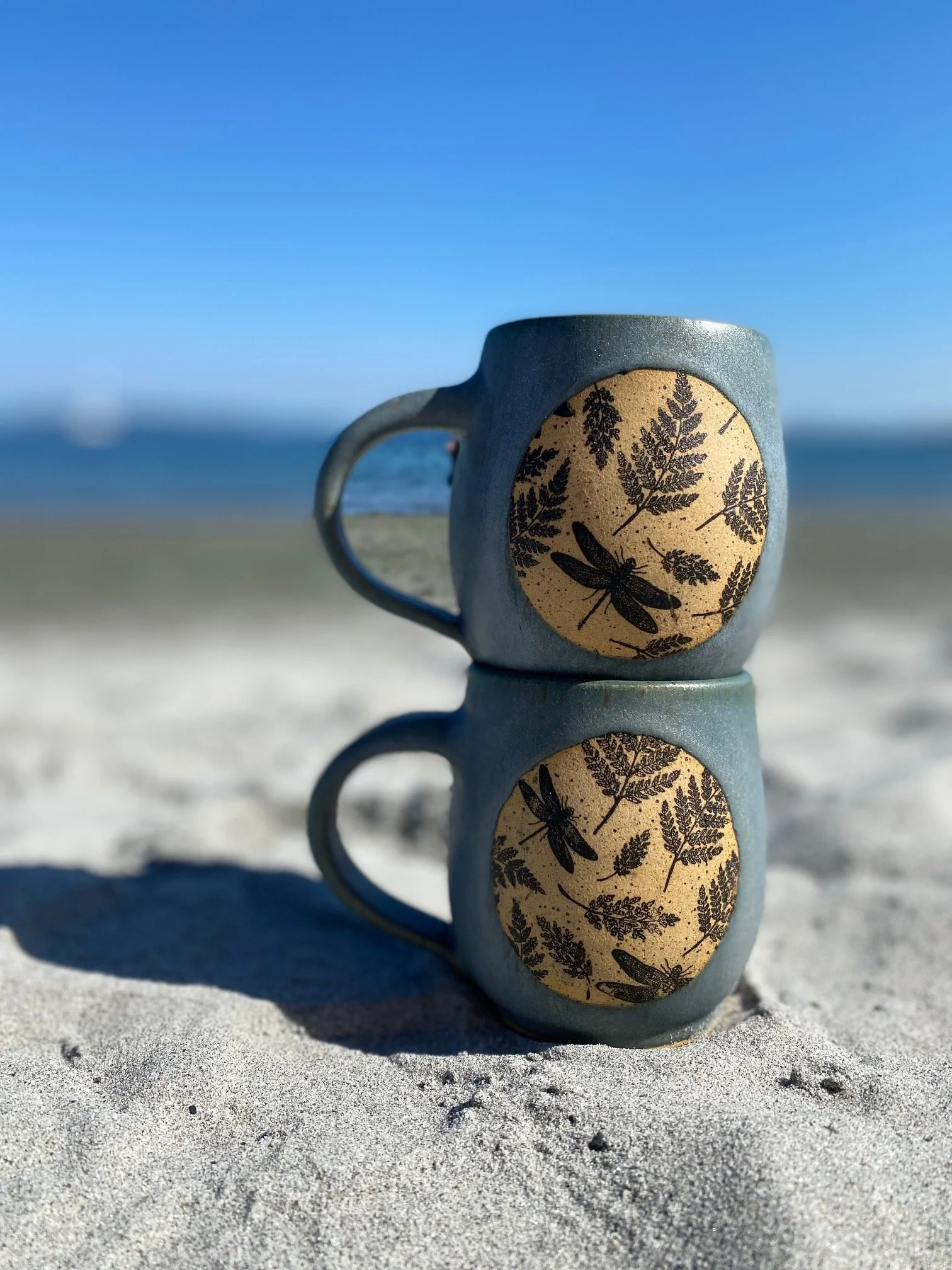 Two ceramic mugs stacked on top of each other on sandy beach, with a blue sky and ocean in the background, each mug has a circular design with black leaves and dragonflies. Sonia Lesage Ceramics