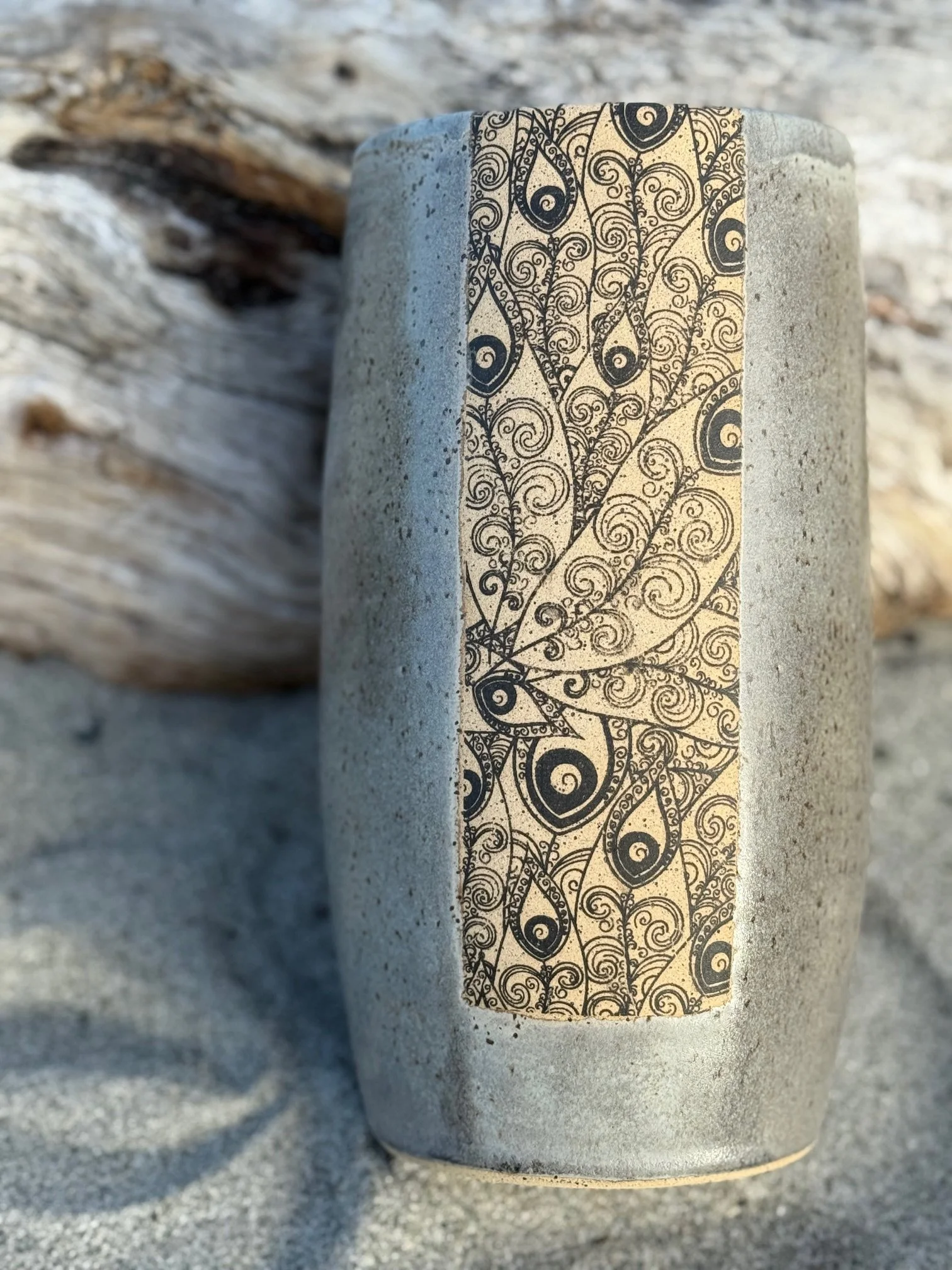 A gray/brown vase on sand with a decorative underglaze transfer on it, with a piece of driftwood in the background. Sonia Lesage Ceramics