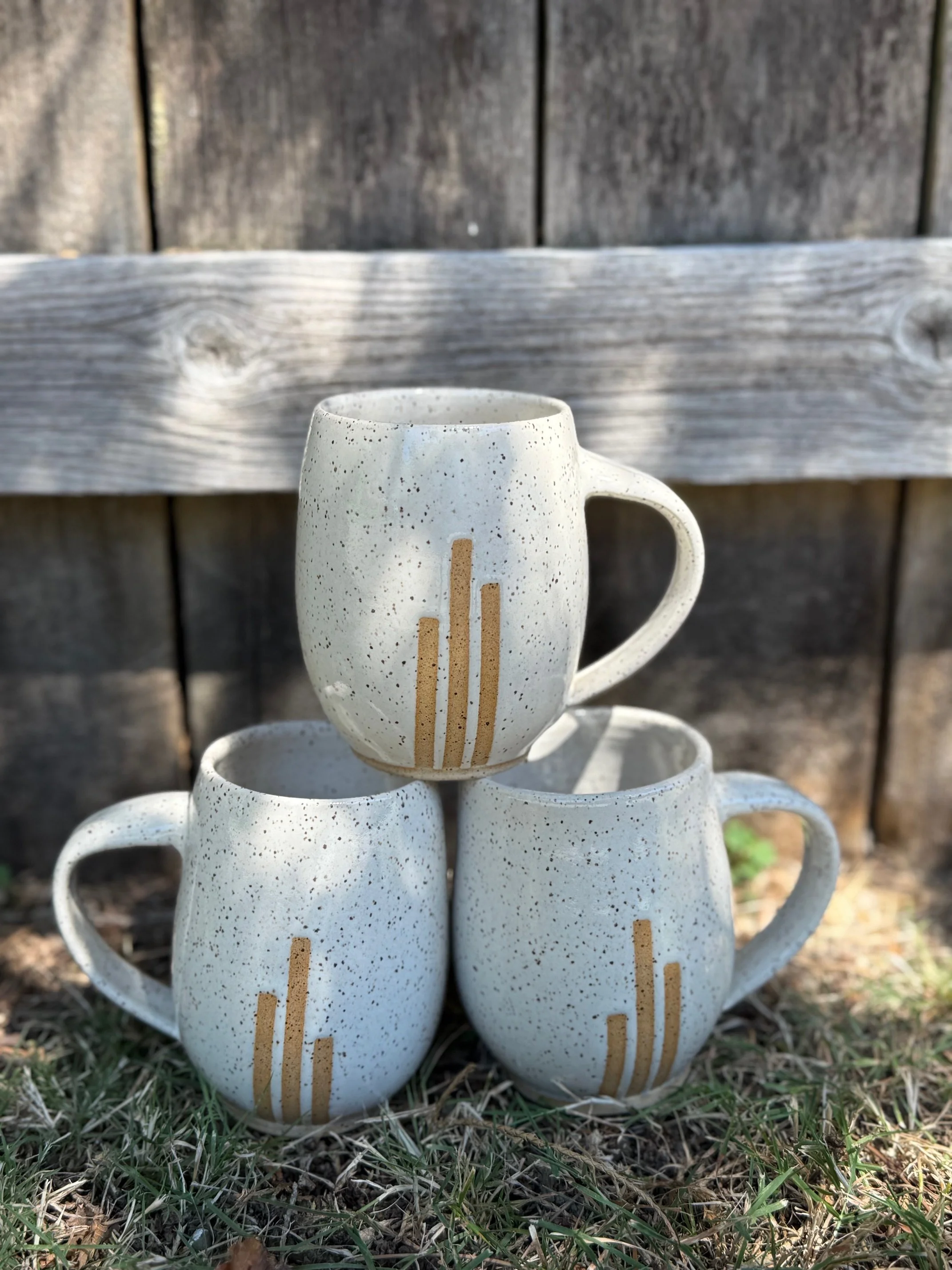 Three ceramic mugs with a speckled white glaze and lines of exposed clay, arranged outdoors against a weathered wooden background, with grass and dirt on the ground. Sonia Lesage Ceramics
