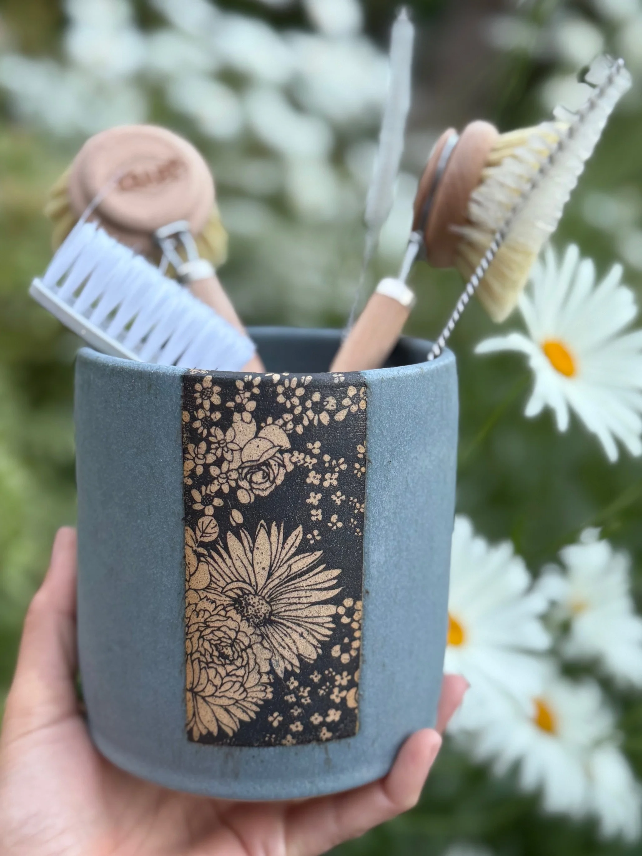 A person's hand holding a ceramic dishbrush vase with a floral design, containing cleaning brushes, with white daisies and greenery in the background. Sonia Lesage Ceramics