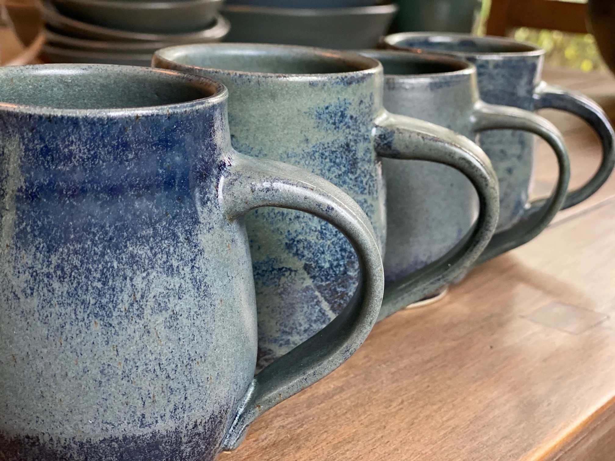 Close-up view of four ceramic coffee mugs with textured blue-gray glaze, arranged on a wooden surface, with some stacked dishes in the background. Sonia Lesage Ceramics