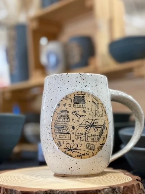 A ceramic mug with speckled and white glaze and a round design with books. Sonia Lesage Ceramics