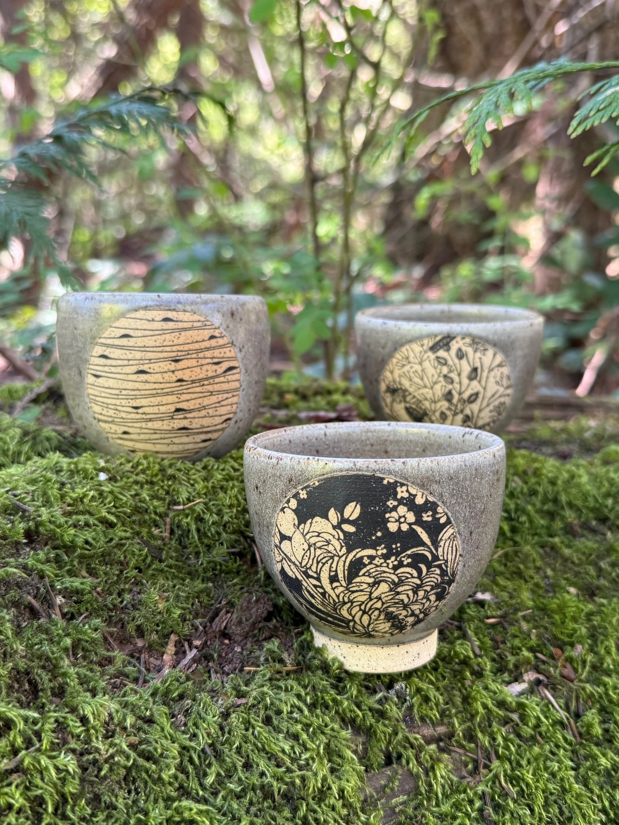Three ceramic espresso/tea cups with black and beige artistic designs, sitting on green moss in a natural woodland setting with plants and trees in the background. Sonia Lesage Ceramics