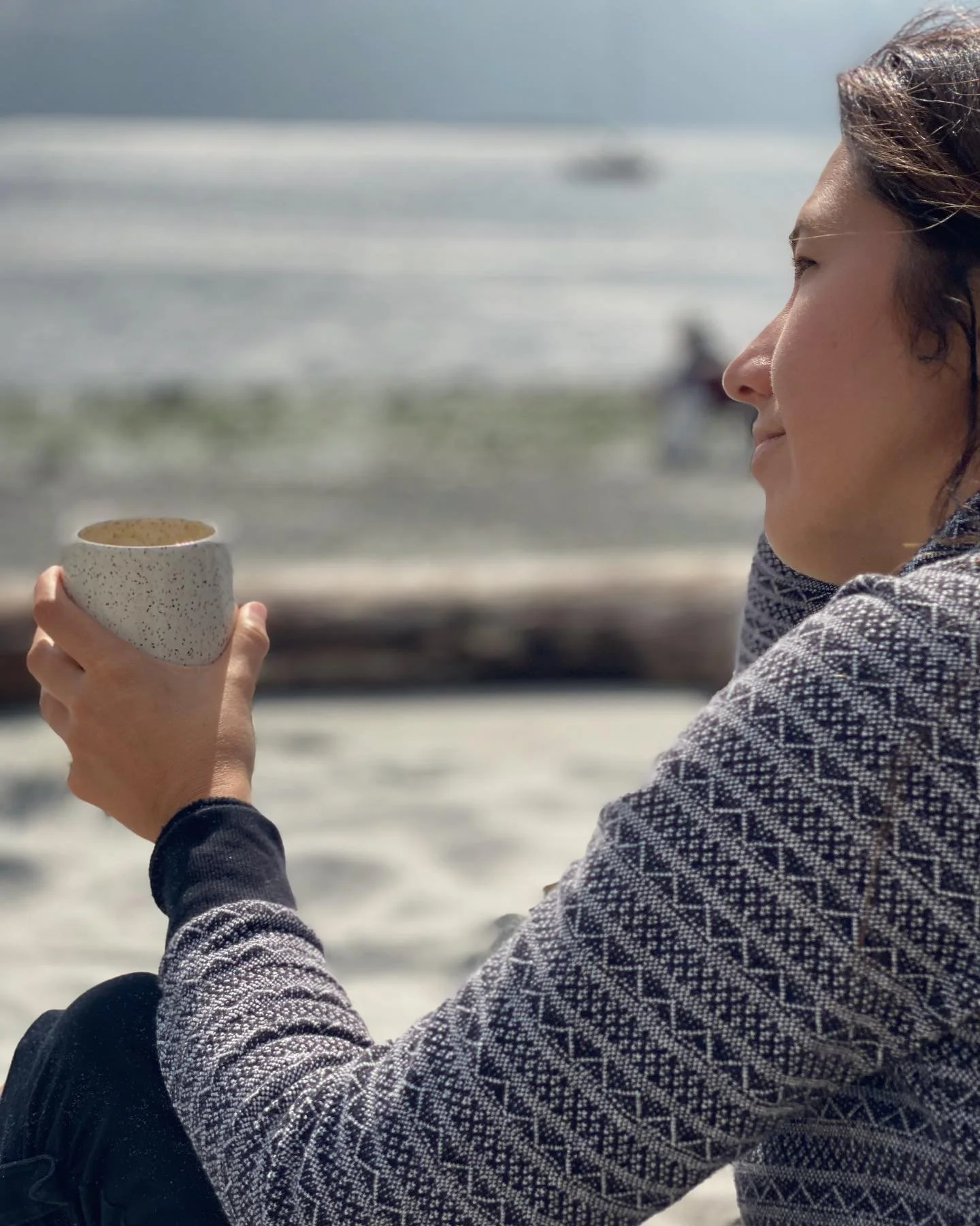 Woman (Sonia Lesage) sitting on the beach, holding a cup of coffee, ocean waves in the background. Sonia Lesage Ceramics