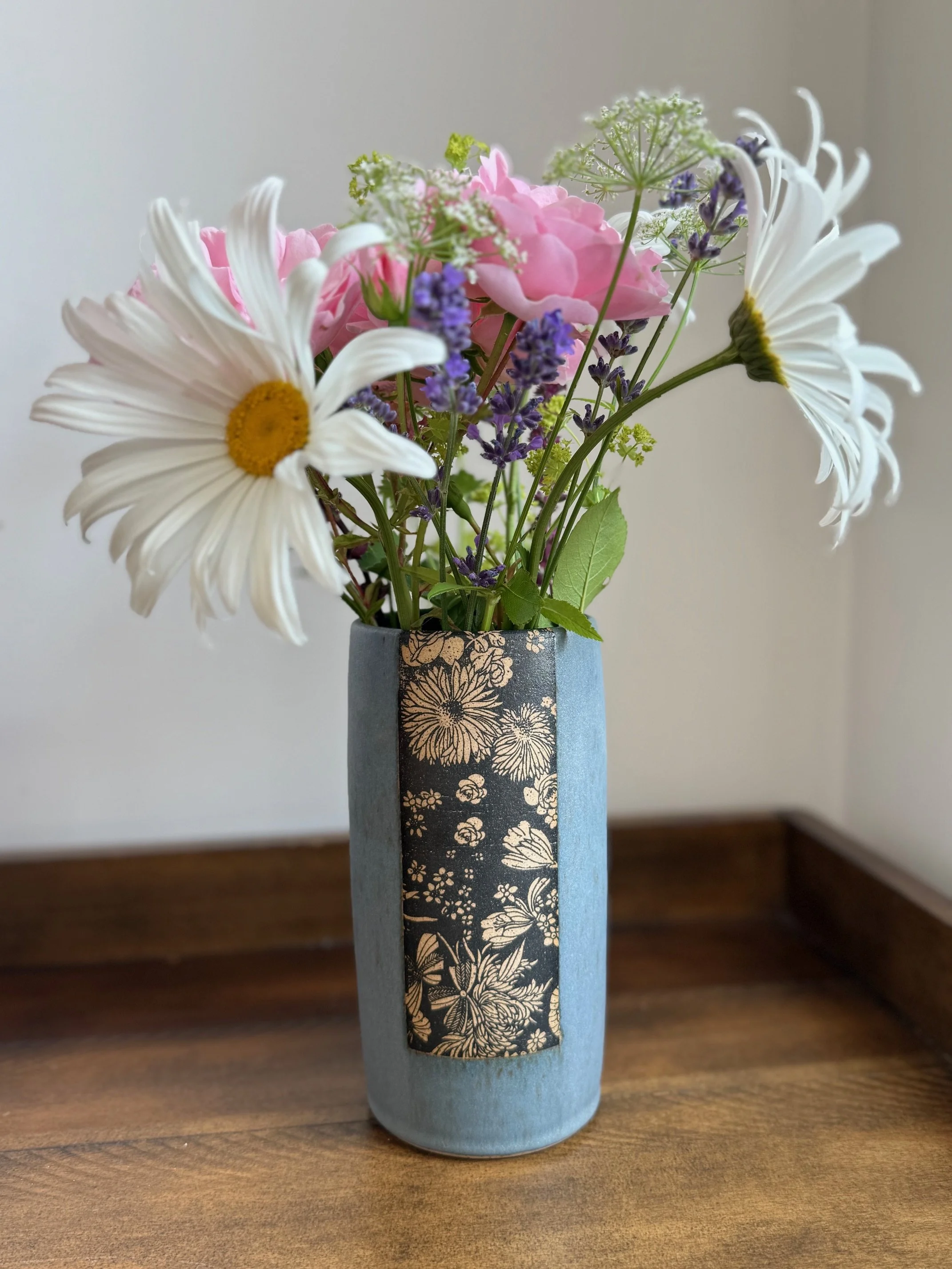 A bouquet of flowers in a blue patterned vase on a wooden surface, consisting of white daisies, pink hydrangeas, purple lavender, and green foliage. Sonia Lesage Ceramics